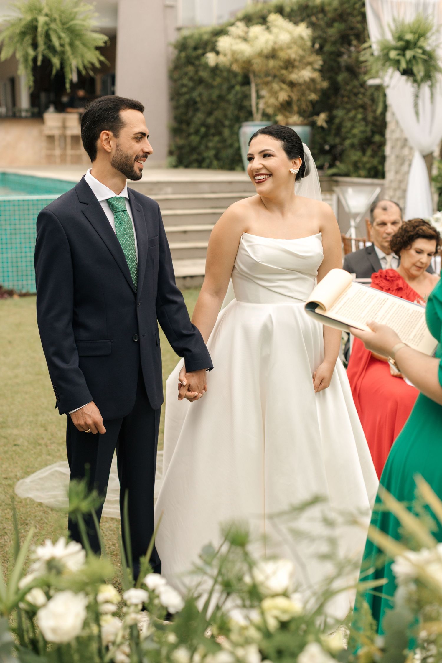 Couple stands together during outdoor garden wedding ceremony beside swimming pool.