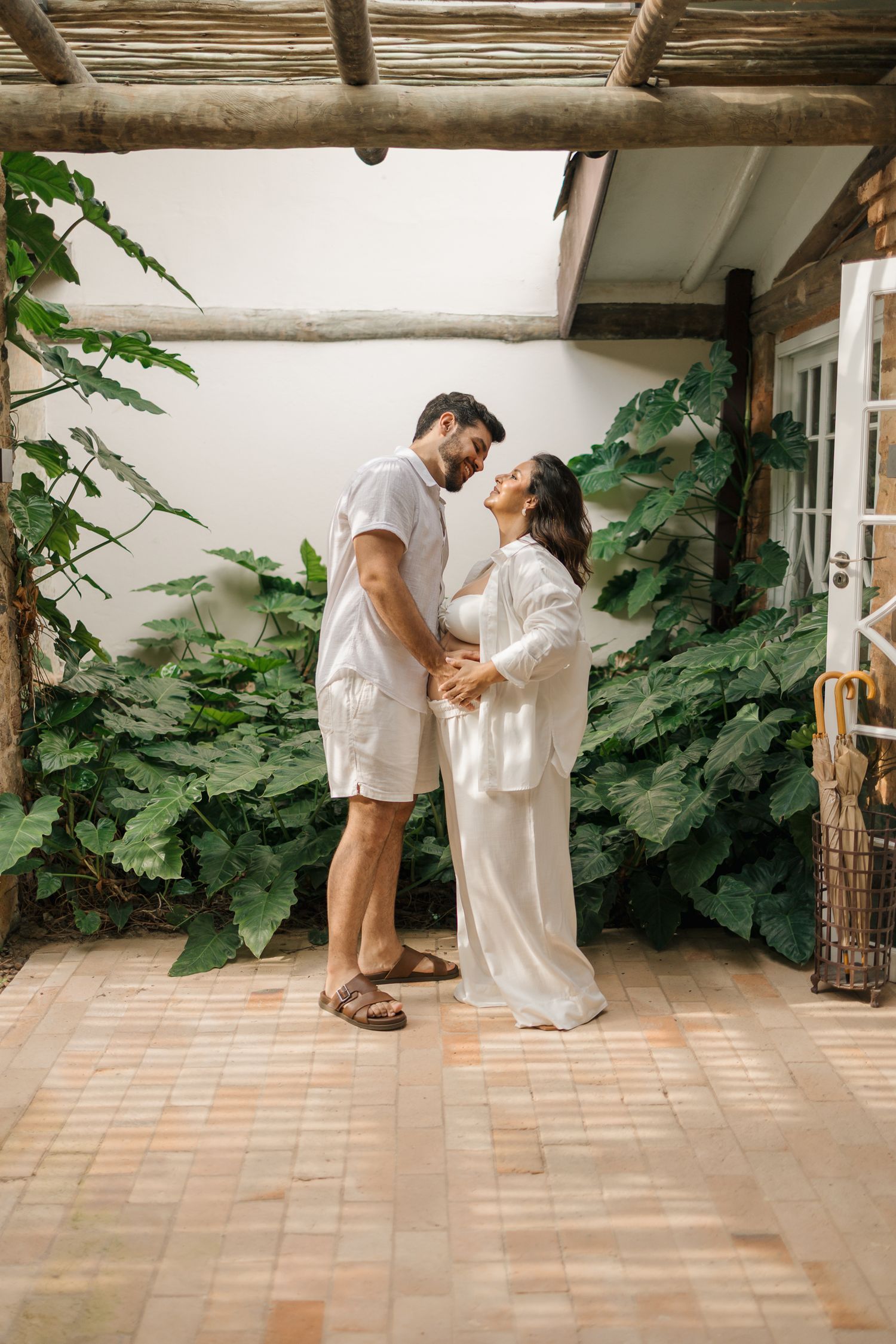 Two people in white clothing share an intimate moment on a tiled patio with lush green plants in the background.