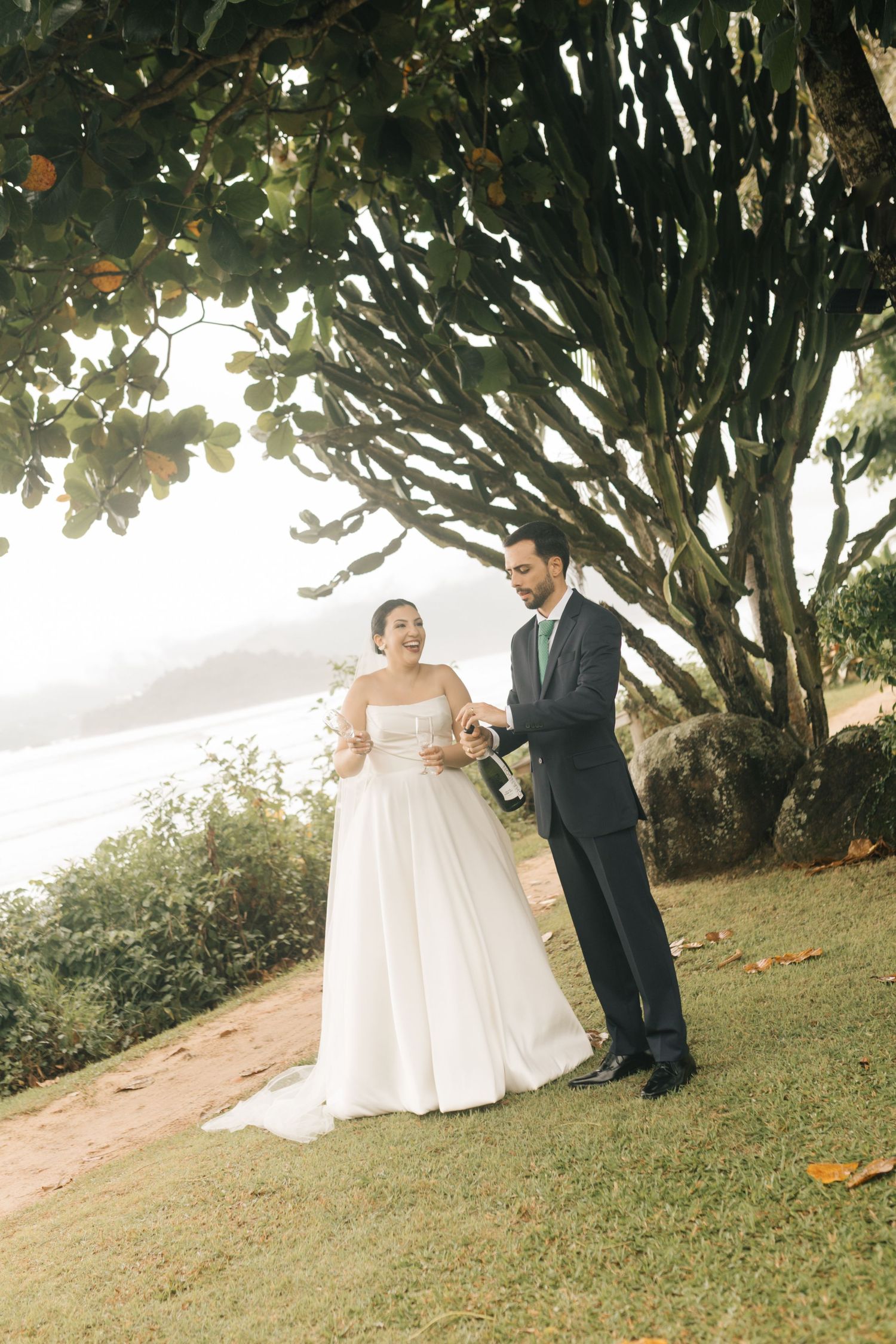 Bride and groom share a joyful moment on a grassy cliff overlooking the ocean.