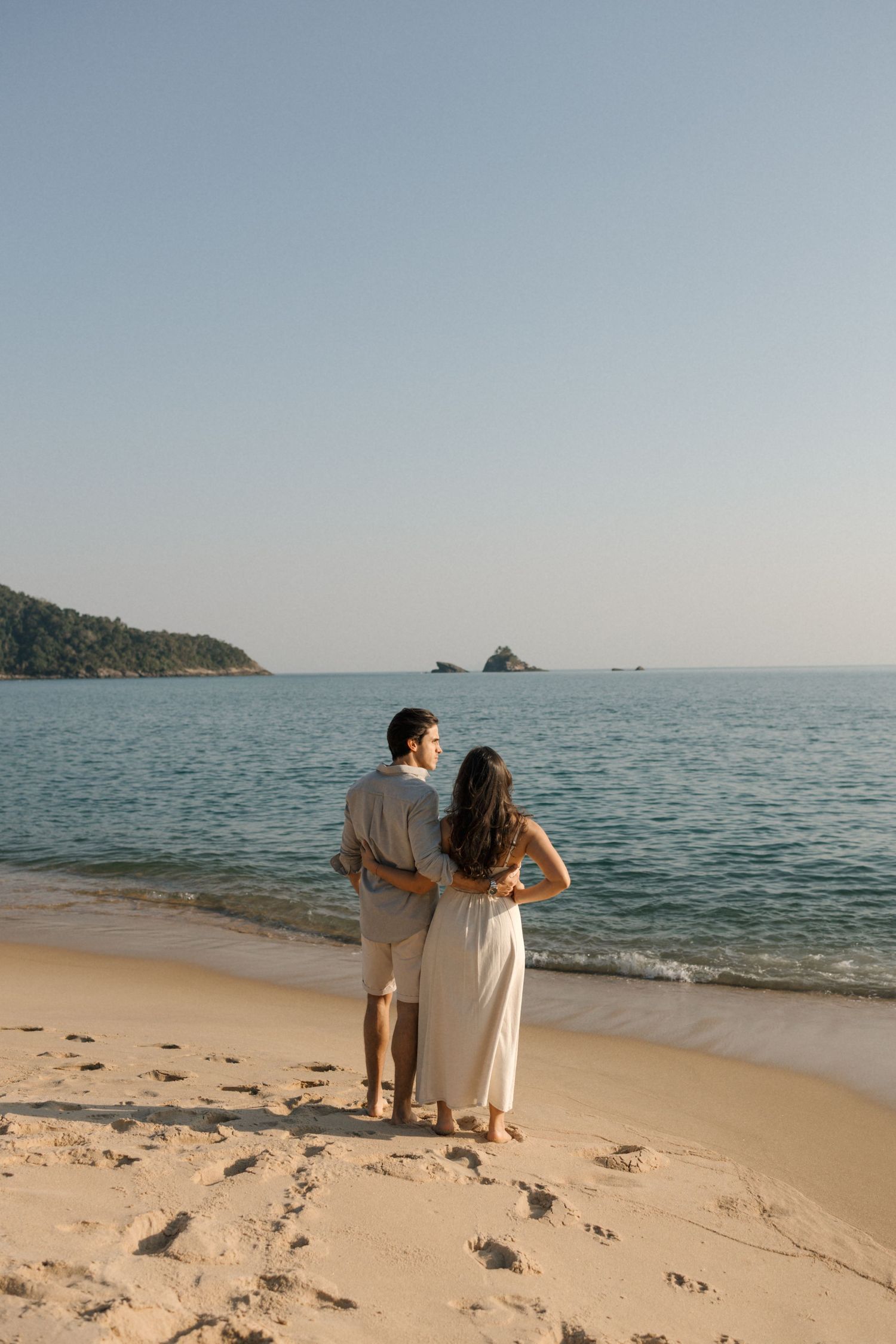 Couple walking together on a sandy beach with calm ocean waters and clear sky in the background.
