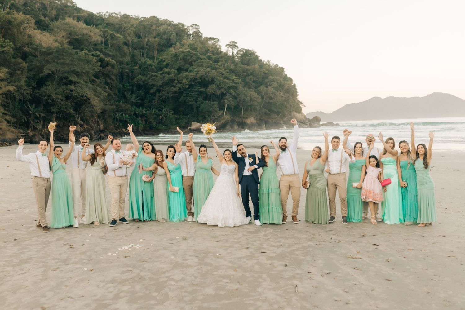 Large wedding party in mint green and neutral dresses posed on a scenic beach.