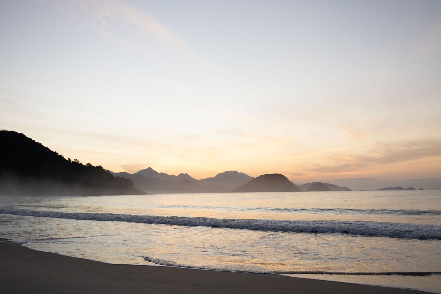 Scenic beach landscape at sunset with mountain silhouettes and gentle waves rolling in.