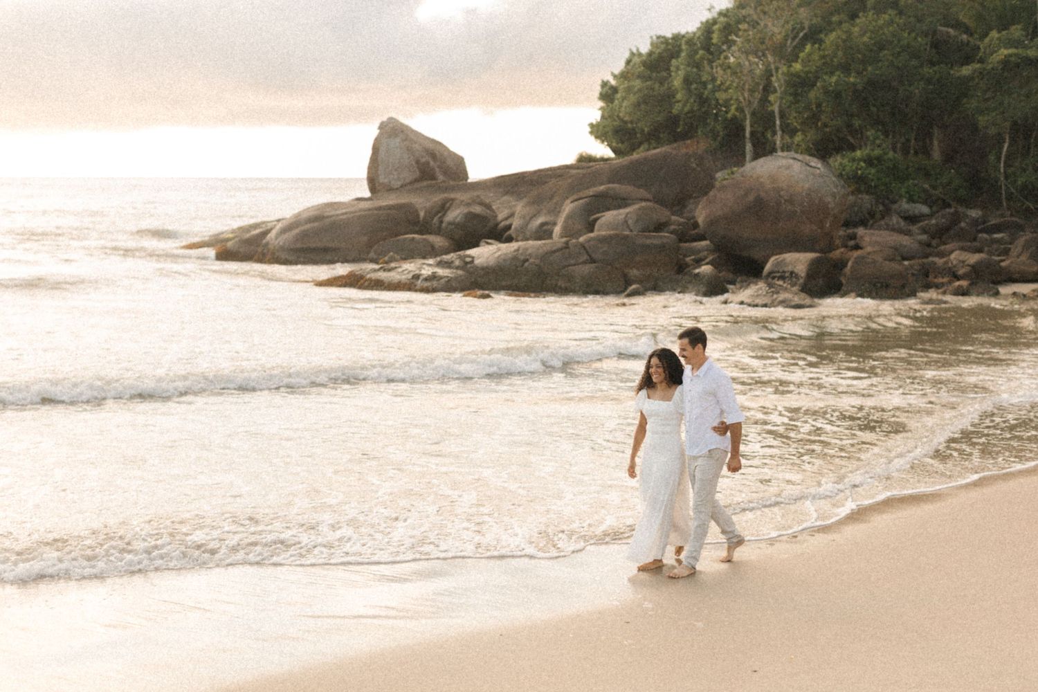 A couple in white clothing takes a romantic sunset walk along a tropical beach with large boulders and ocean waves.