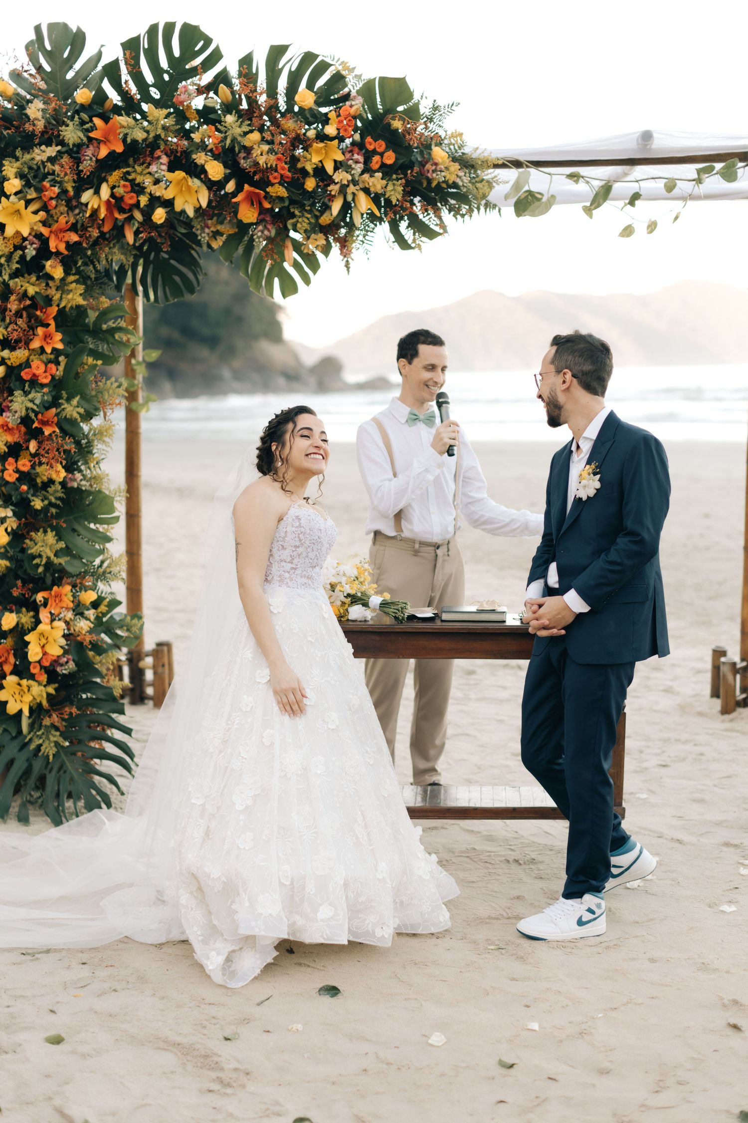 Beach wedding ceremony under an arch decorated with monstera leaves and orange flowers at sunset.