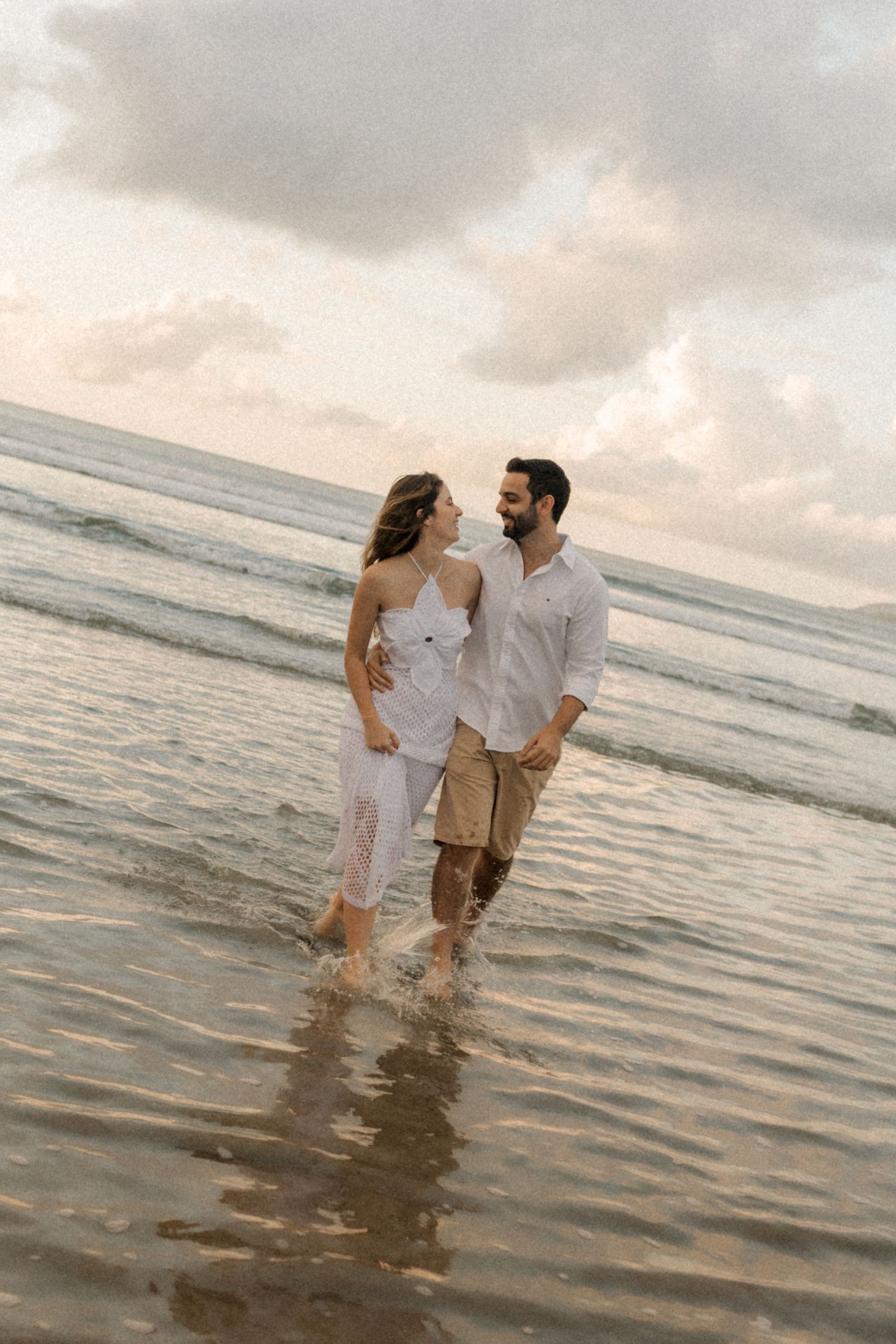 Couple walking together in shallow ocean water during sunset with cloudy sky in the background.