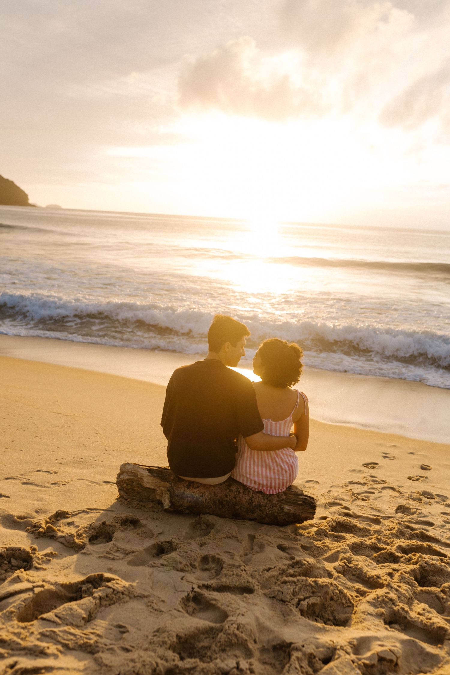 Um casal está sentado em uma rocha observando o pôr do sol na praia enquanto as ondas quebram na costa dourada.