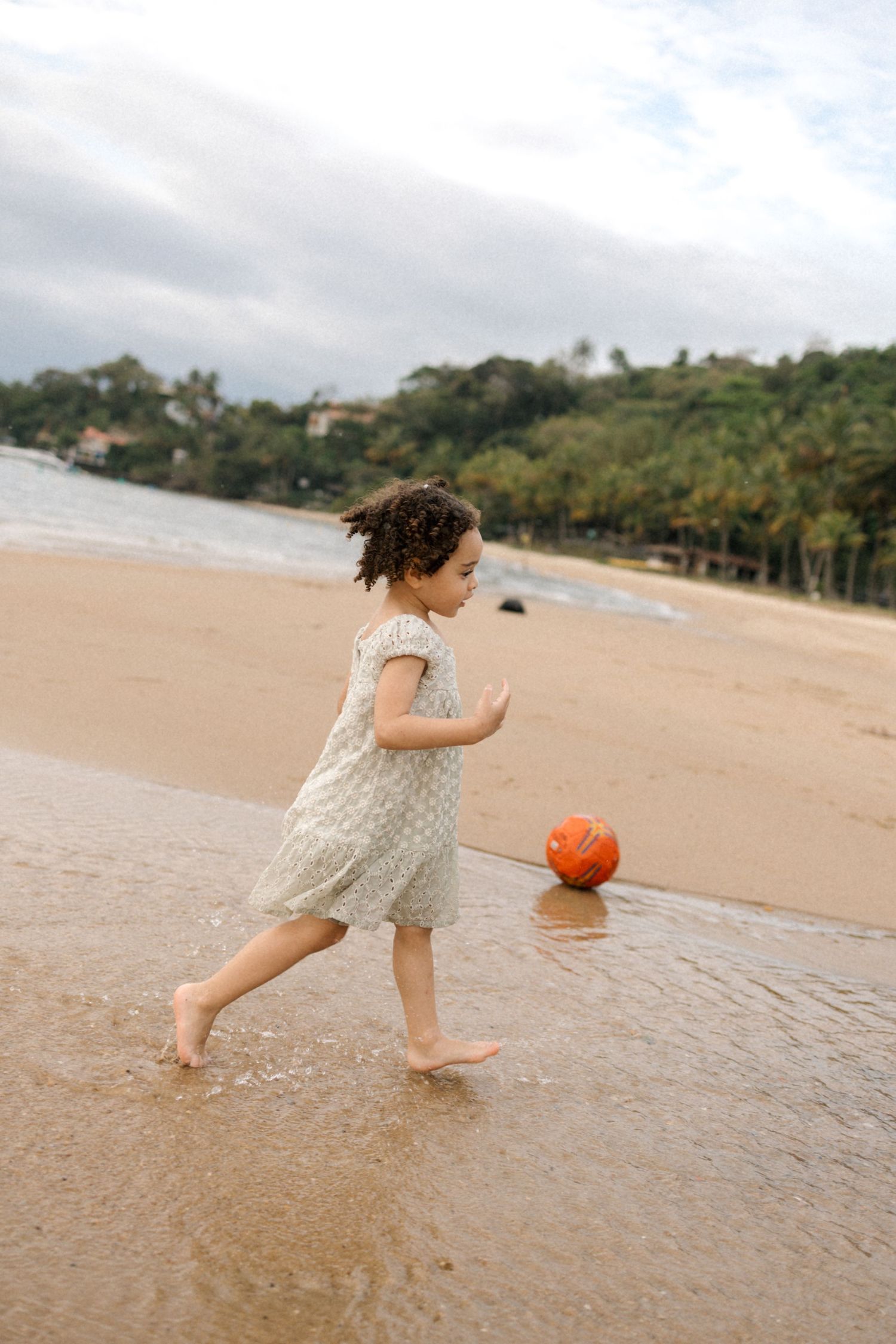 A child in a polka dot dress runs along the shoreline chasing an orange ball on a sandy beach.