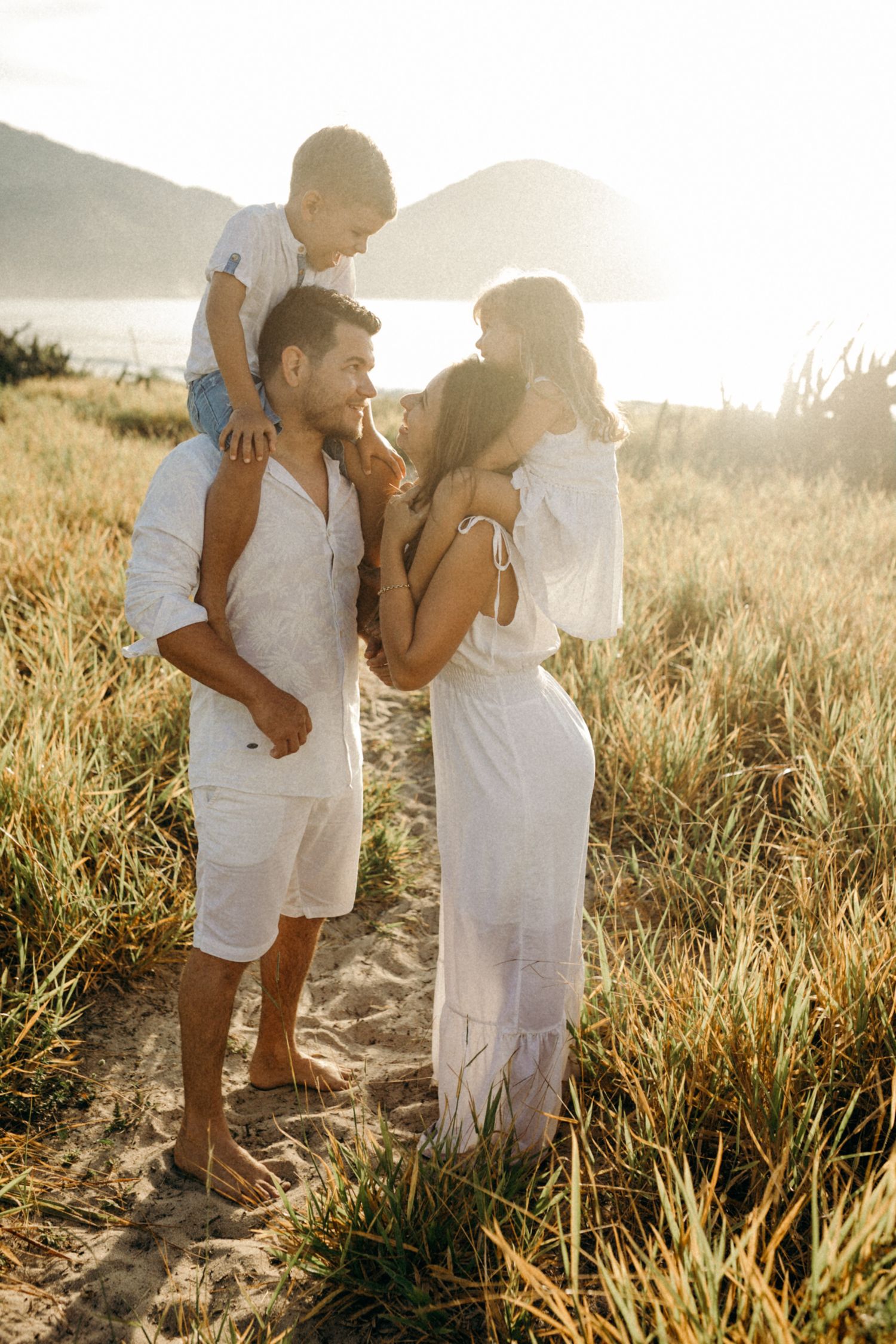Family in white outfits walking together through sunlit grassy field at sunset.