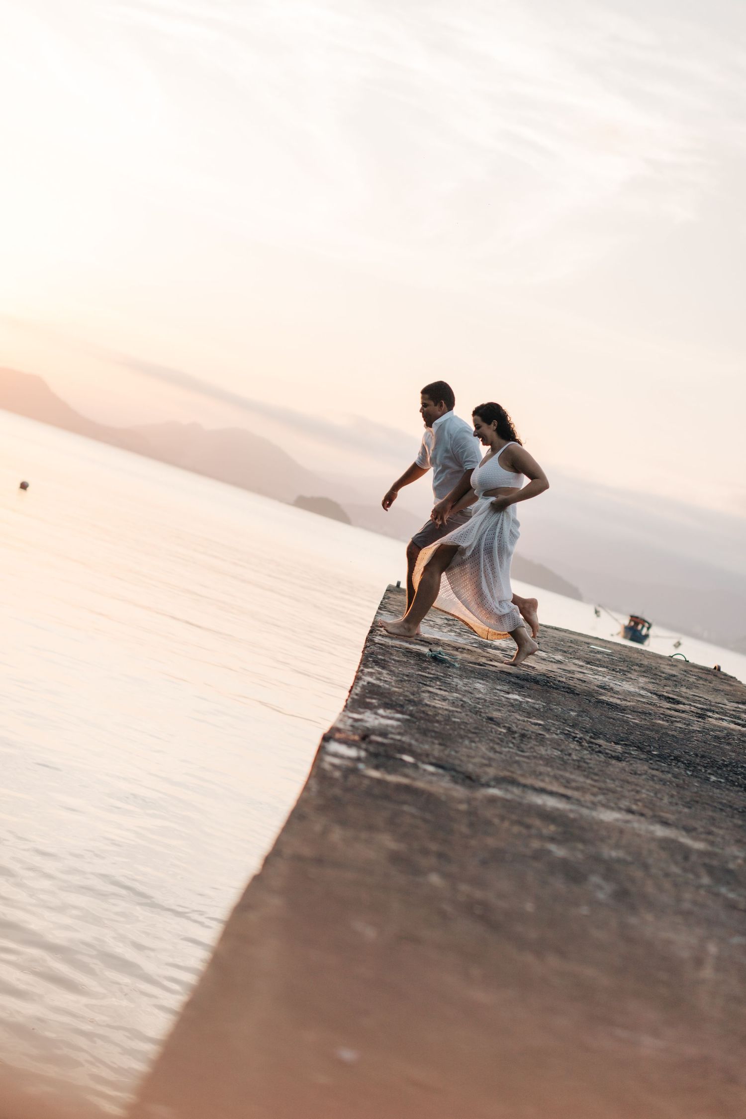 A couple runs playfully along a concrete pier during sunset by the water.