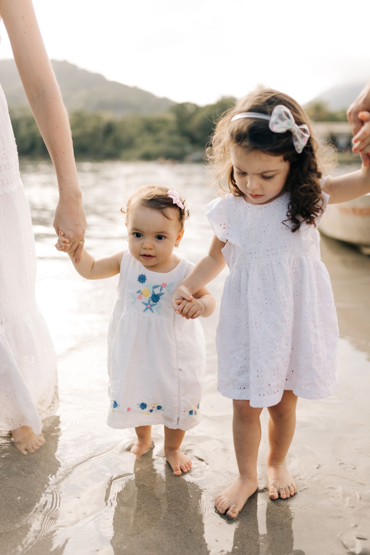 Two children in white dresses holding hands while walking along beach shore.