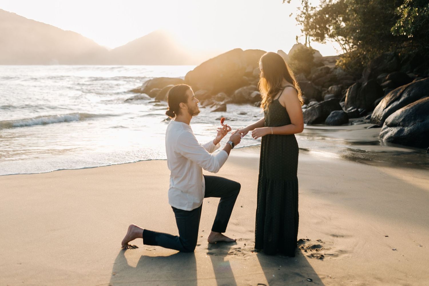 Emotional beach proposal moment captured during sunset with rocky coastline backdrop.