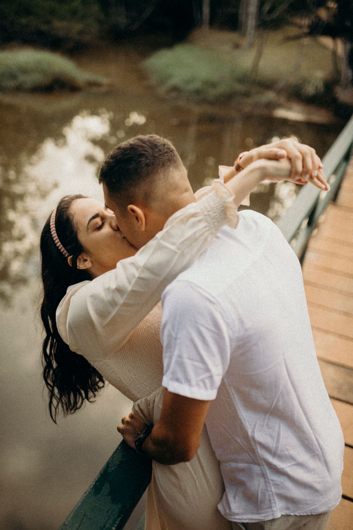 Romantic couple embracing and kissing on a wooden bridge over calm water at sunset.