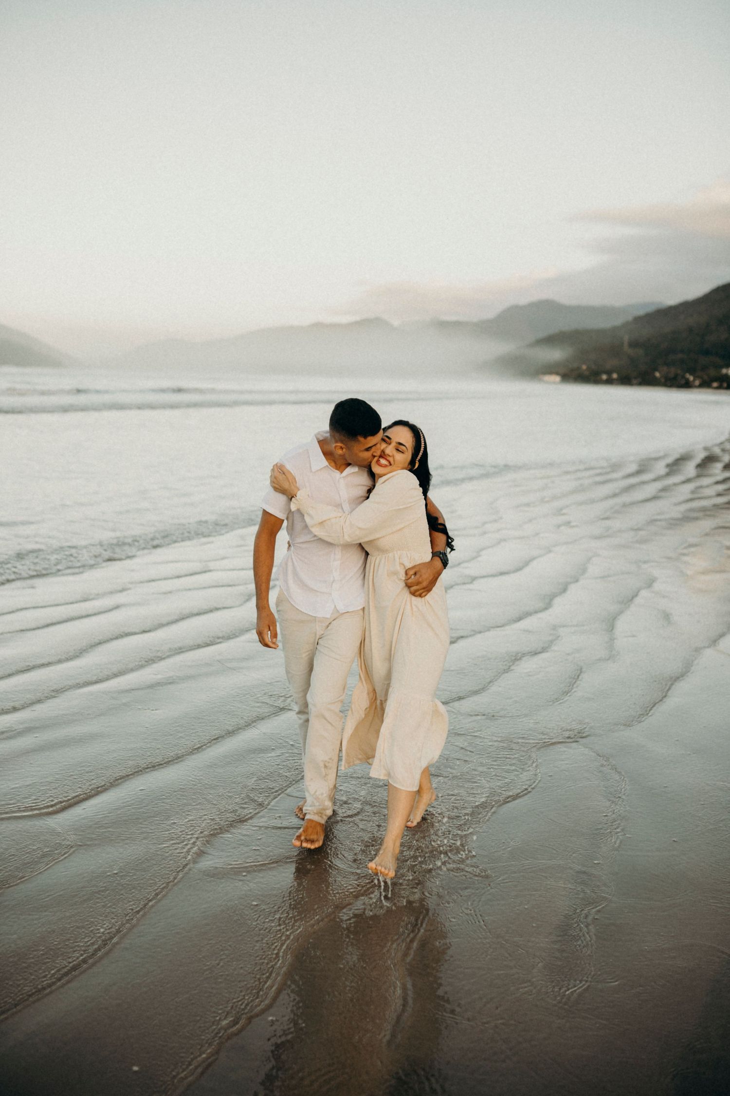 Two people in light colored clothing stroll together on a wet beach with mountains in the distance.
