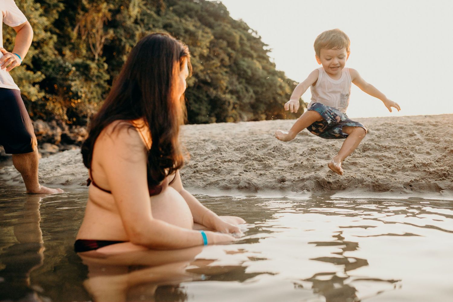 Child jumps with joy in shallow beach water as waves splash near parent on a sunny afternoon.