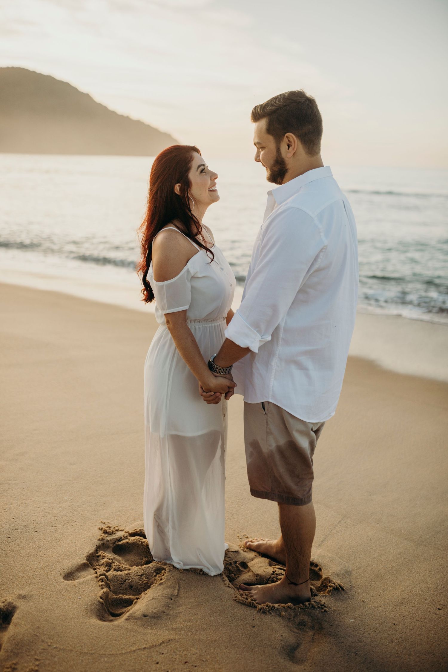 Couple in white clothing gazing at each other on misty beach at sunset.