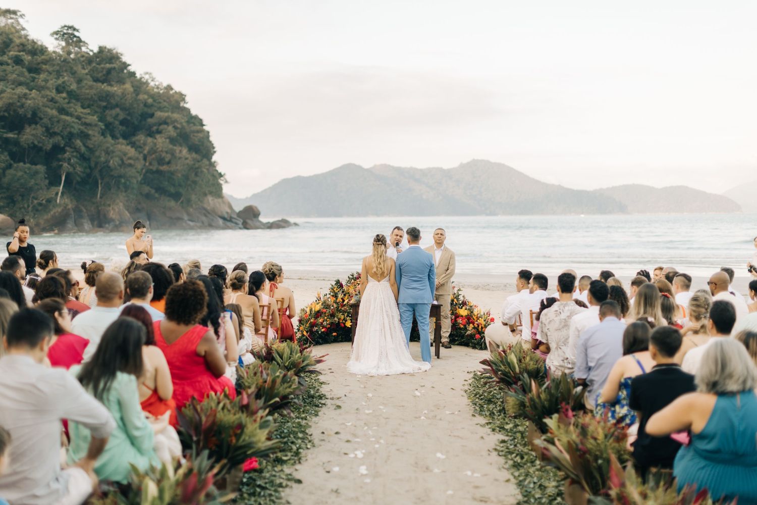 Beach wedding ceremony setup with guests and floral arrangements against coastal mountain backdrop.