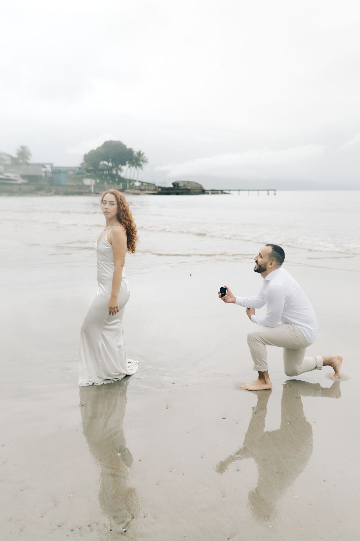 Surprise beach proposal with person kneeling on wet sand while other faces away.