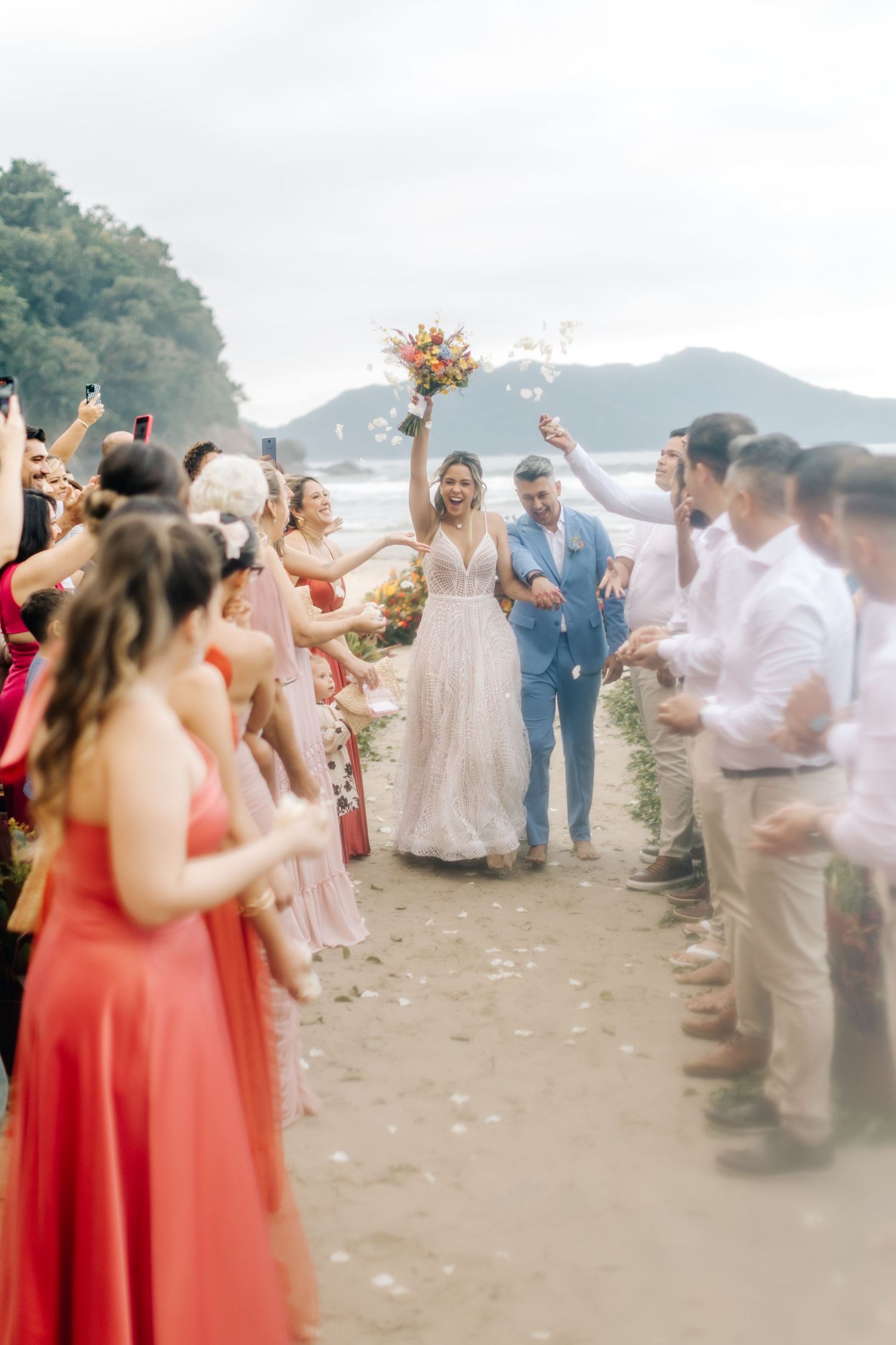 Wedding guests form a celebration line on the beach with coral bridesmaid dresses and white attire.