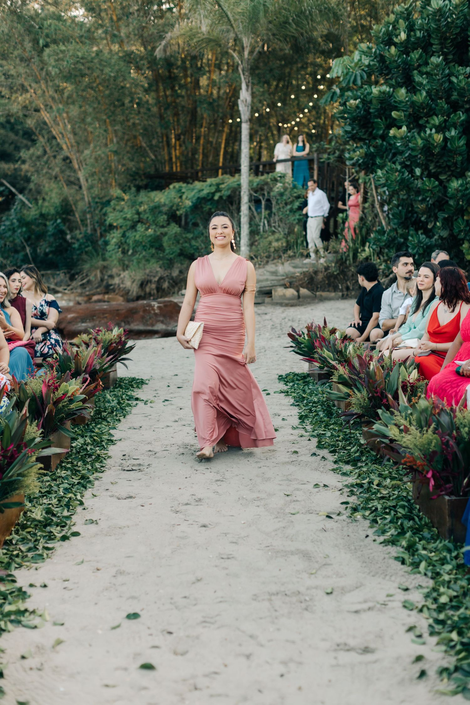 Guest in rose-colored formal dress walks down decorated outdoor aisle.
