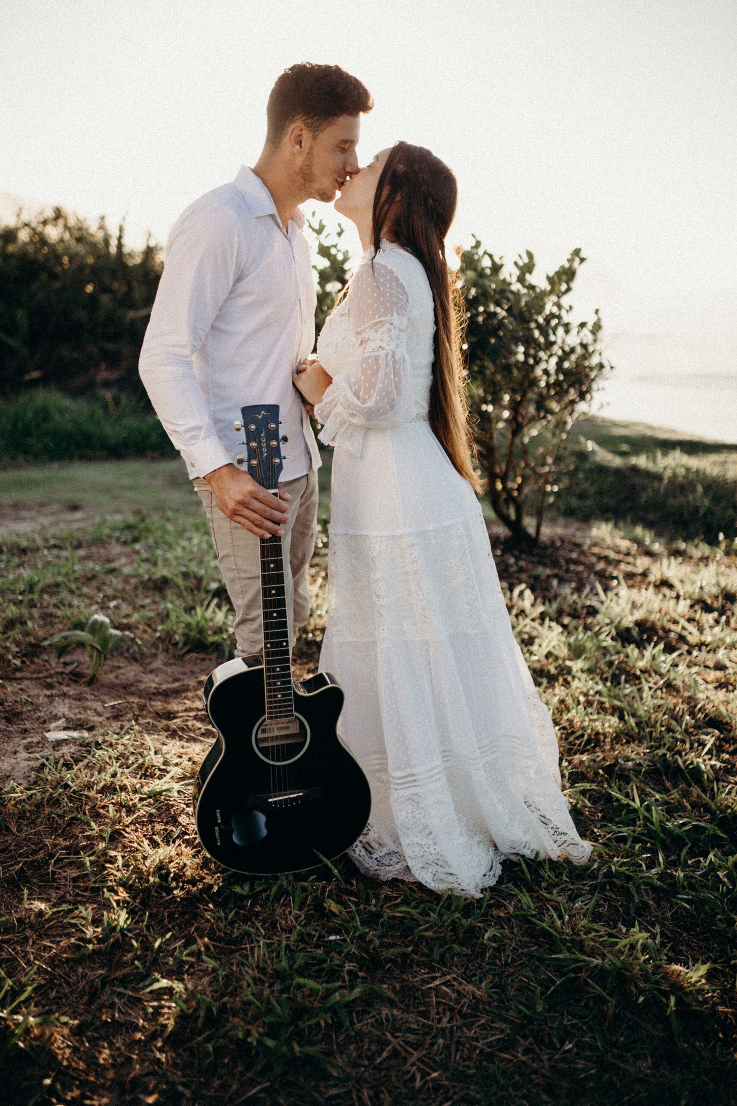 Couple in white wedding attire pose with black guitar against scenic nature backdrop.