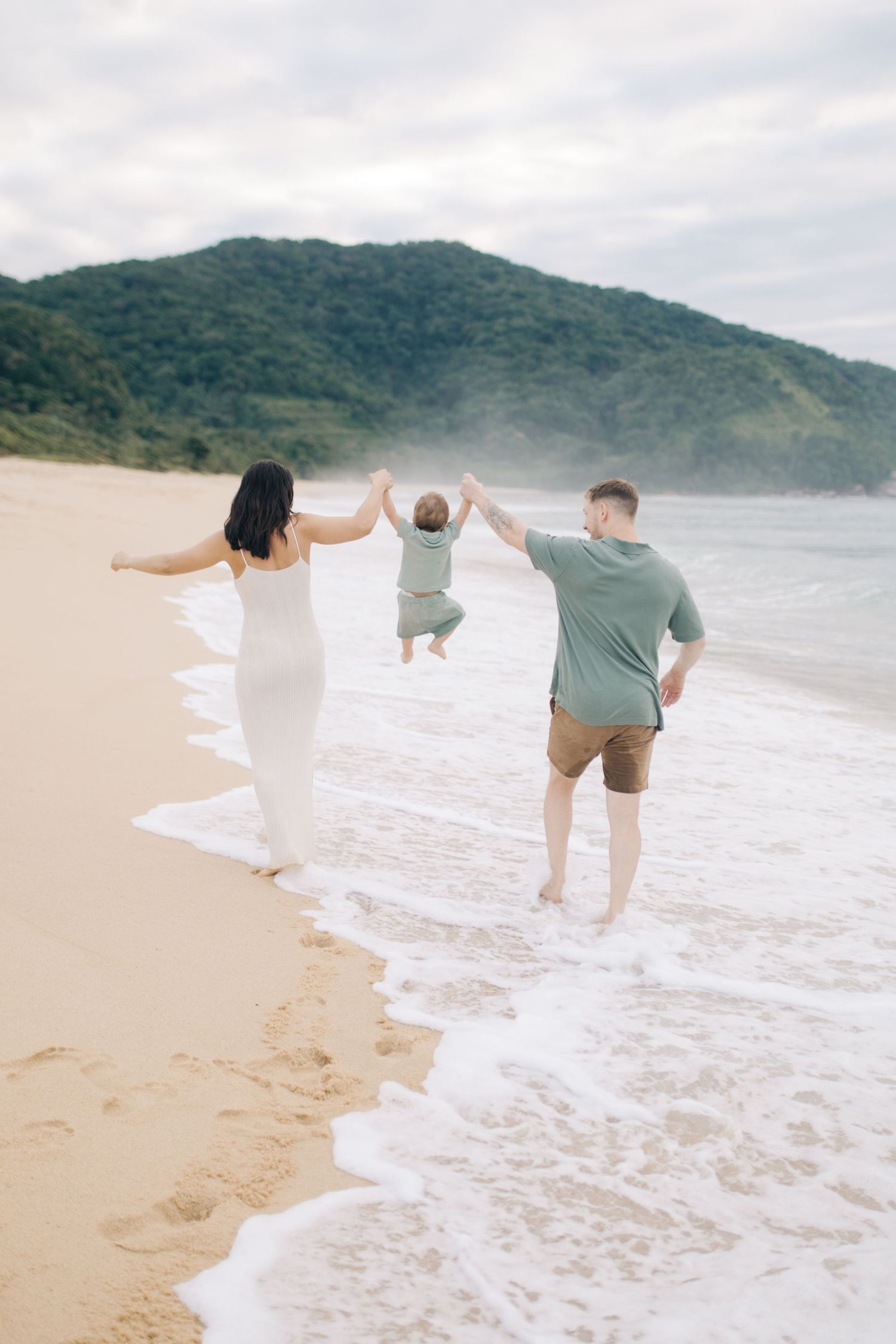 Family holds hands while playing in the ocean waves at a scenic beach surrounded by mountains.
