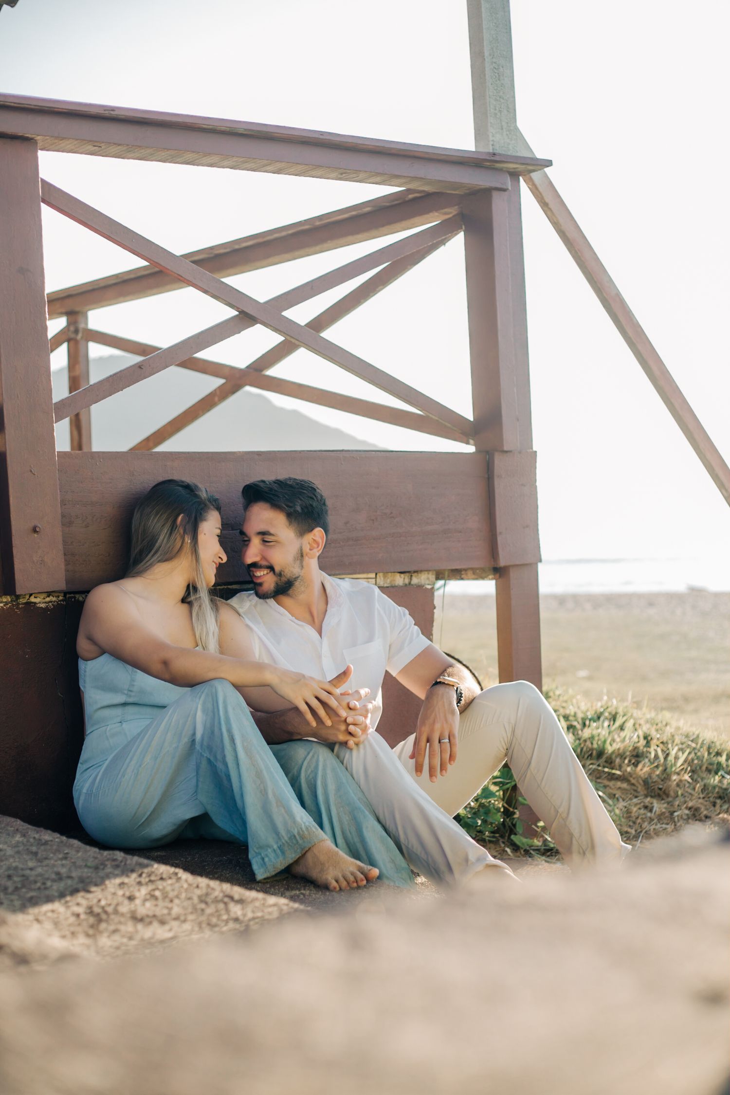 Tender couple in light blue dress and white outfit sitting close together on wooden deck railing.