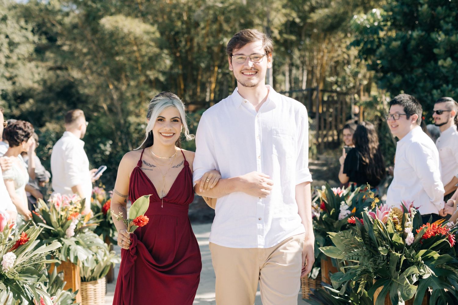 Wedding celebration with guests in burgundy and white attire near tropical arrangements.