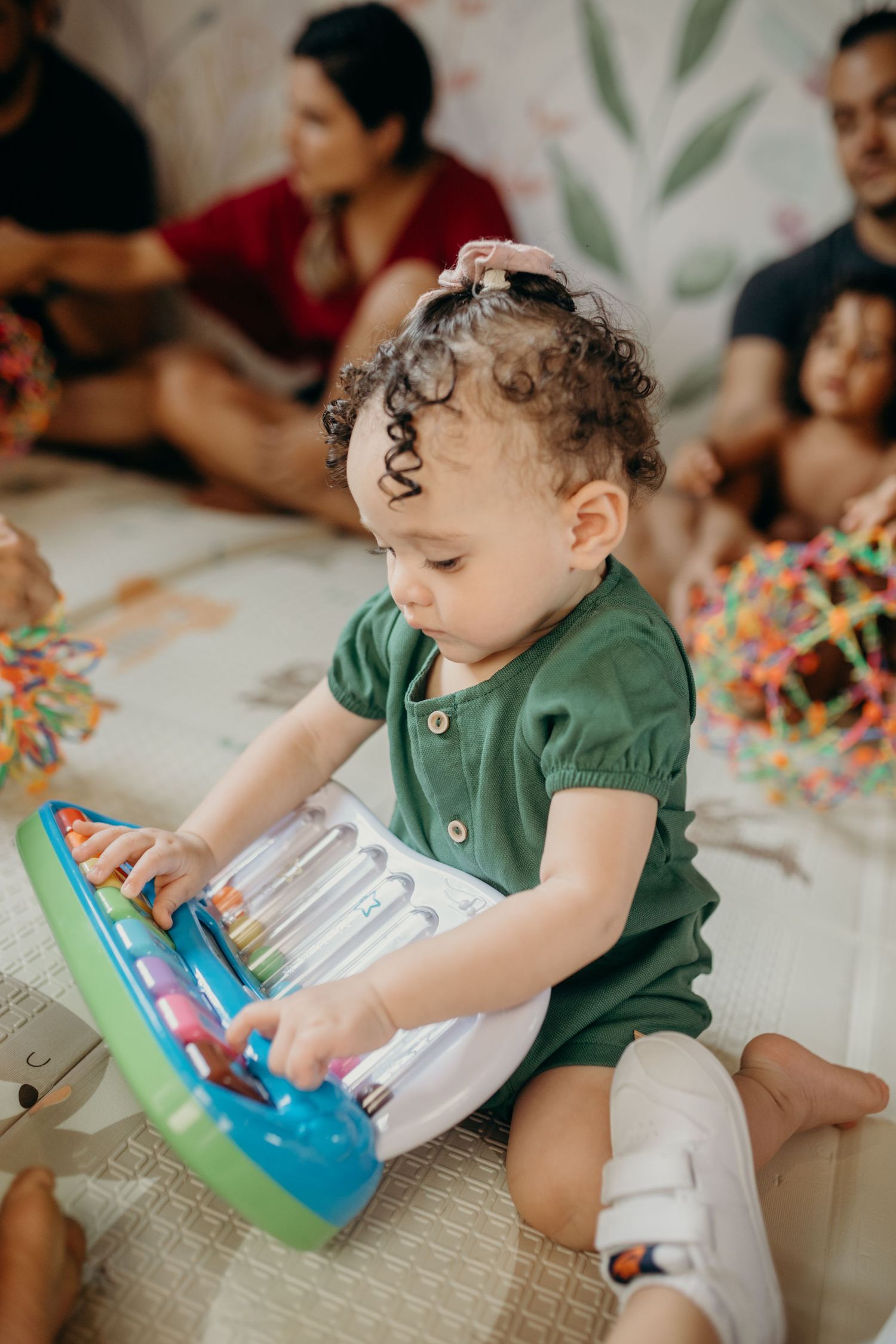 A small child in a green outfit exploring toys while sitting on a patterned mat.
