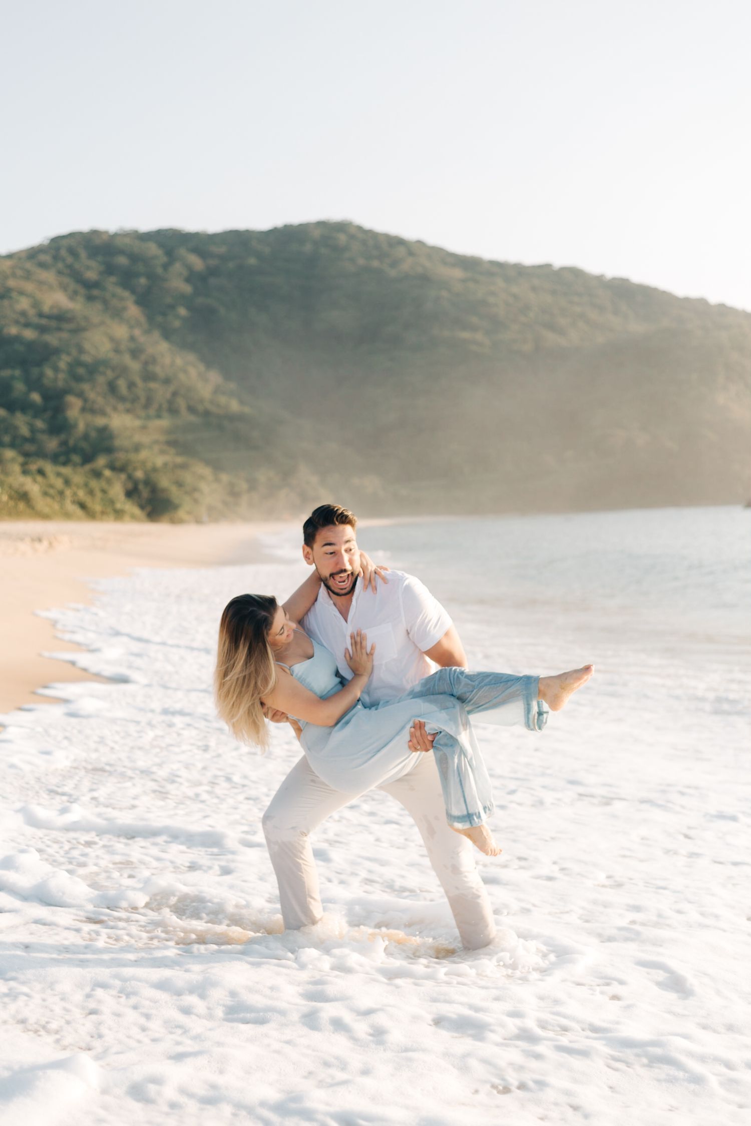 Joyful couple playing and lifting each other on beach with crashing waves.