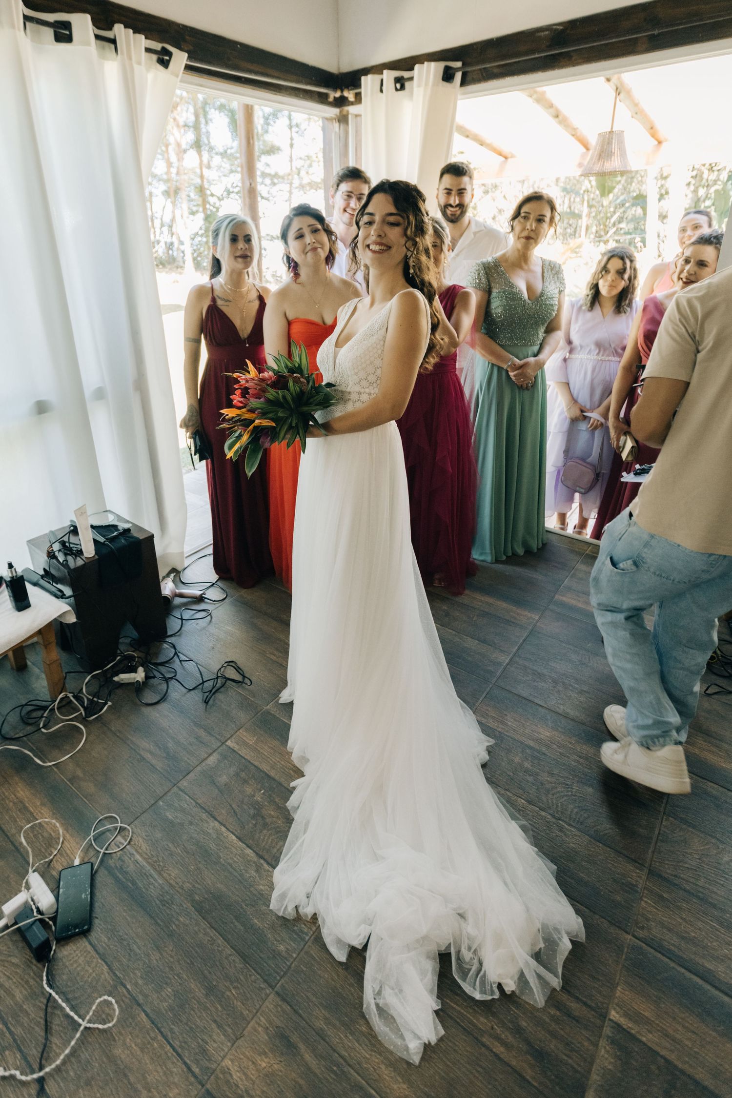 Wedding party gathered in colorful dresses on wooden floors with white curtain backdrop.