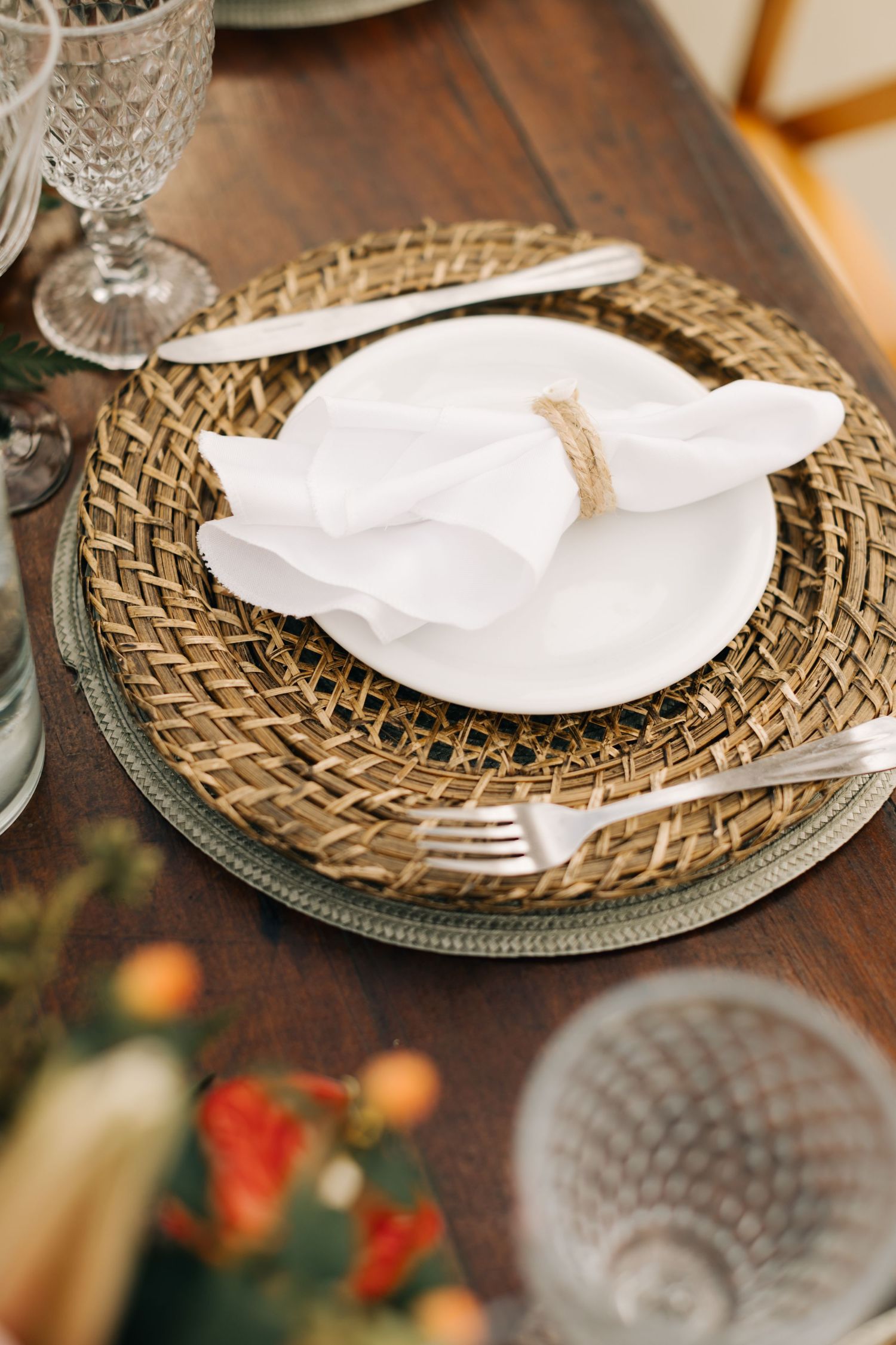Place setting with white napkin on woven charger plate and crystal glassware on wooden table.