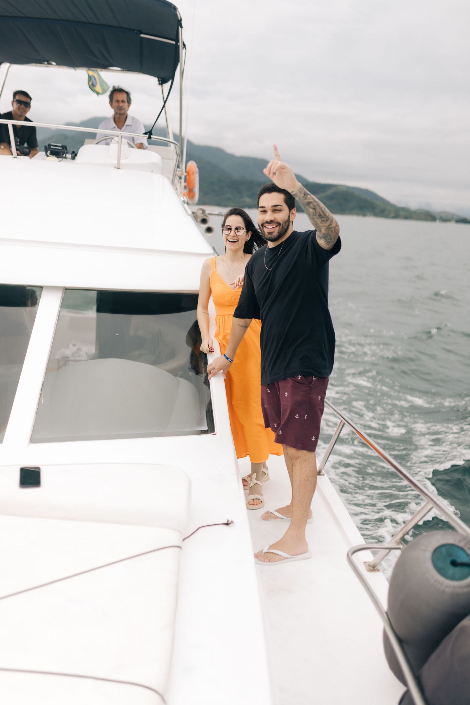 People waving from yacht deck with mountain landscape behind them.