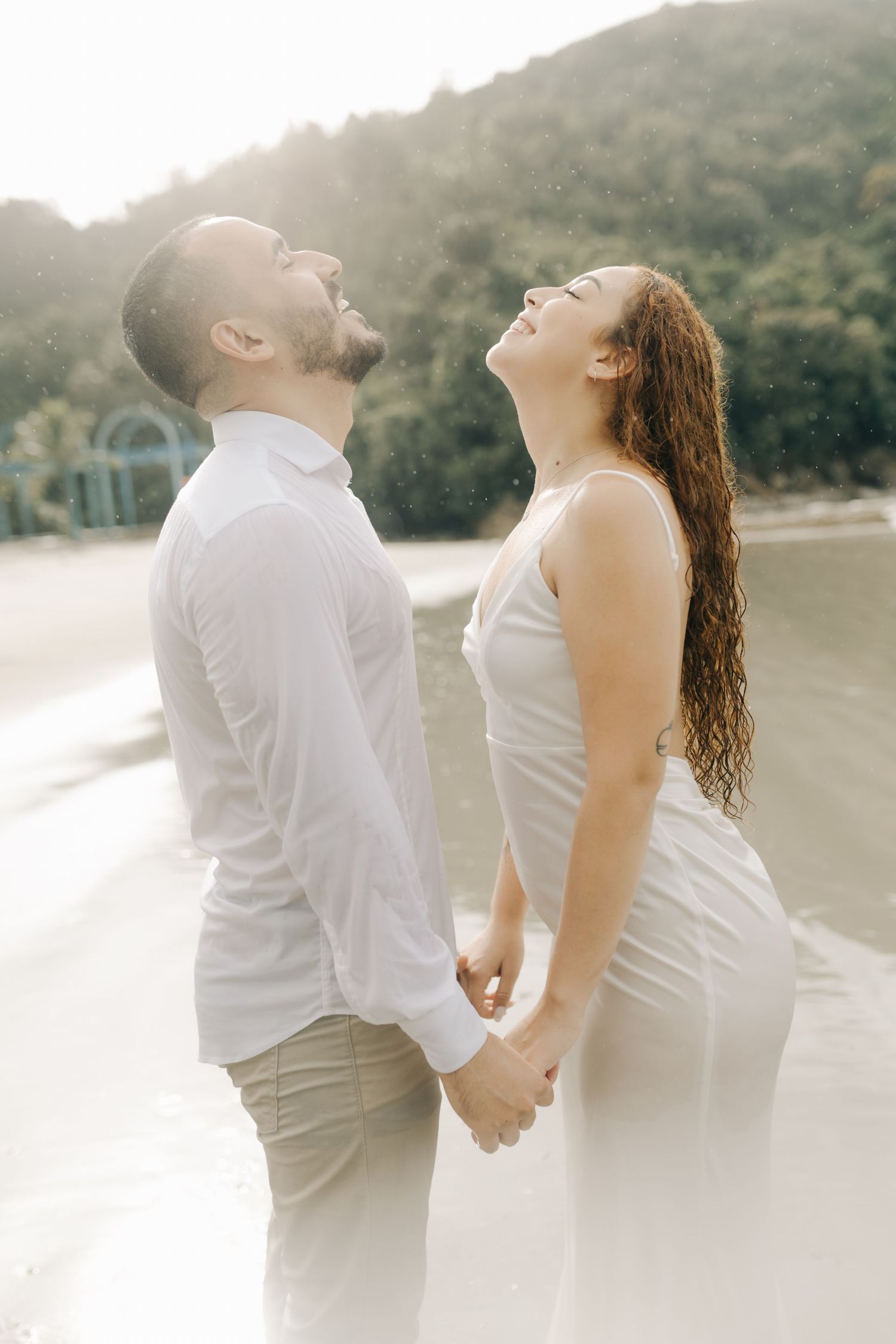 Couple in white clothing dancing and laughing together on a serene beach.