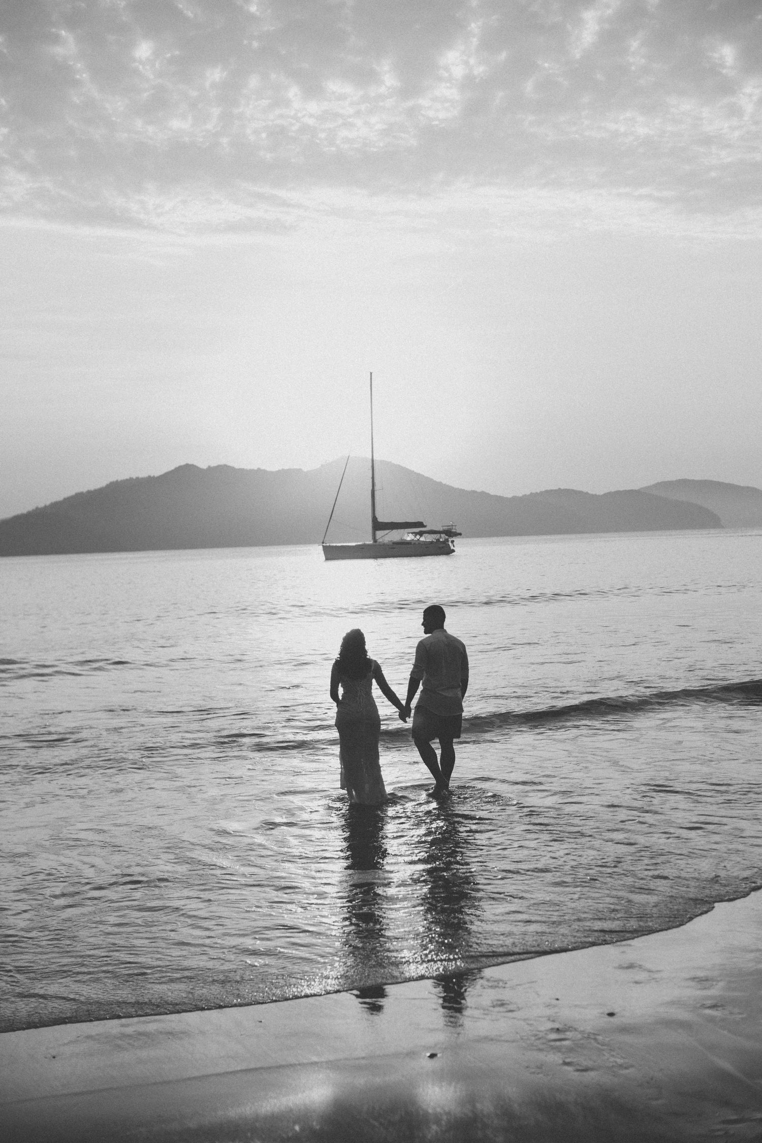 Silhouetted couple stands in ocean waves with sailboat and mountains in black and white.