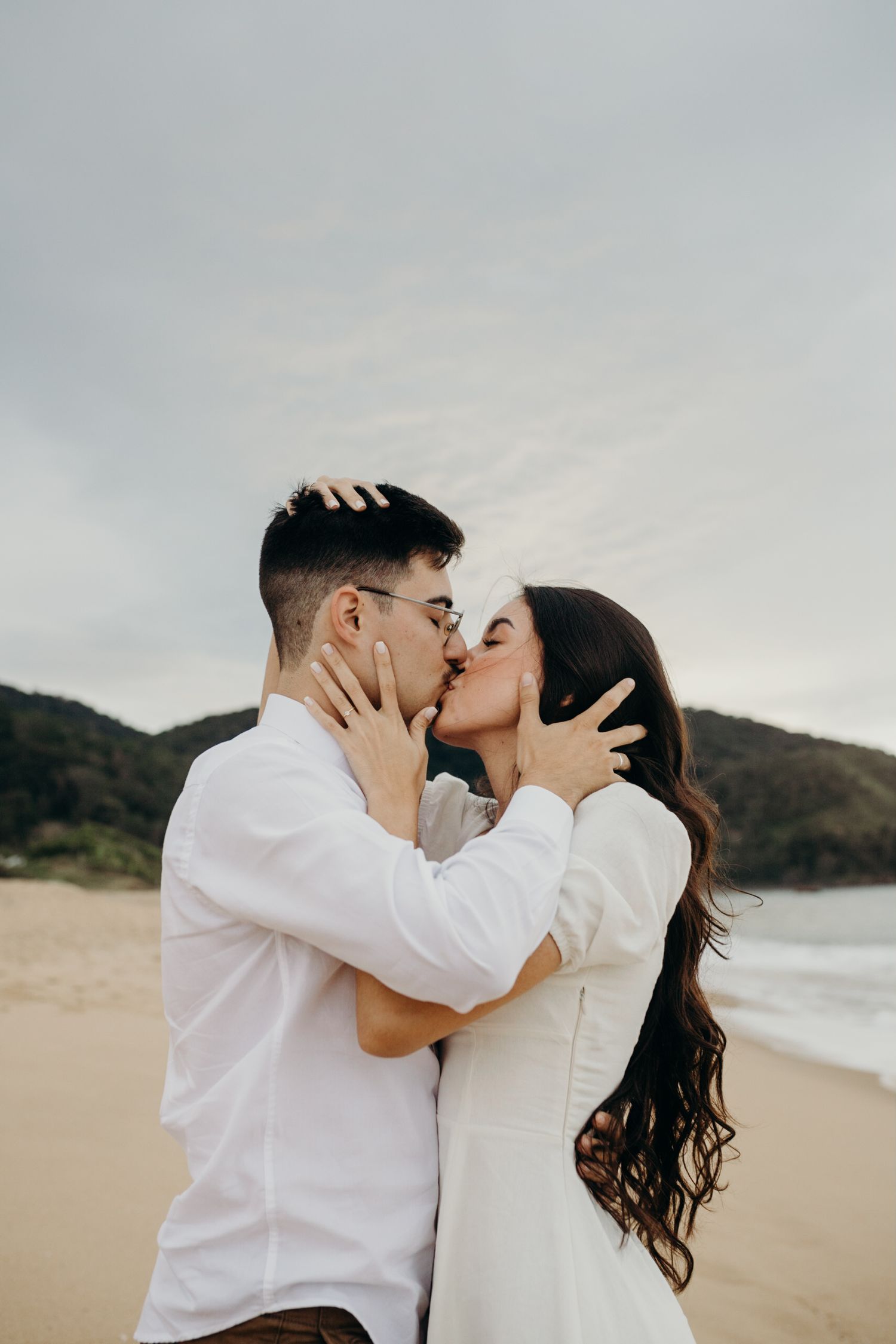 A passionate embrace captured on a scenic beach with hills in the background.