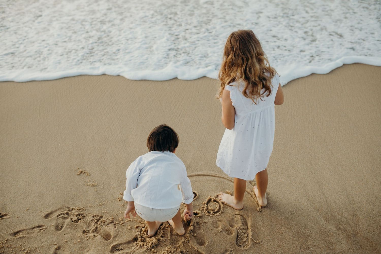 Children drawing in the wet sand near ocean waves at sunset.