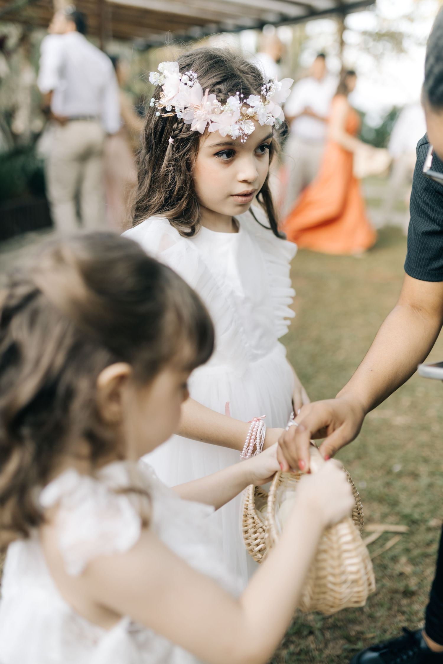 Young attendants in white dresses carry baskets at an outdoor wedding ceremony.