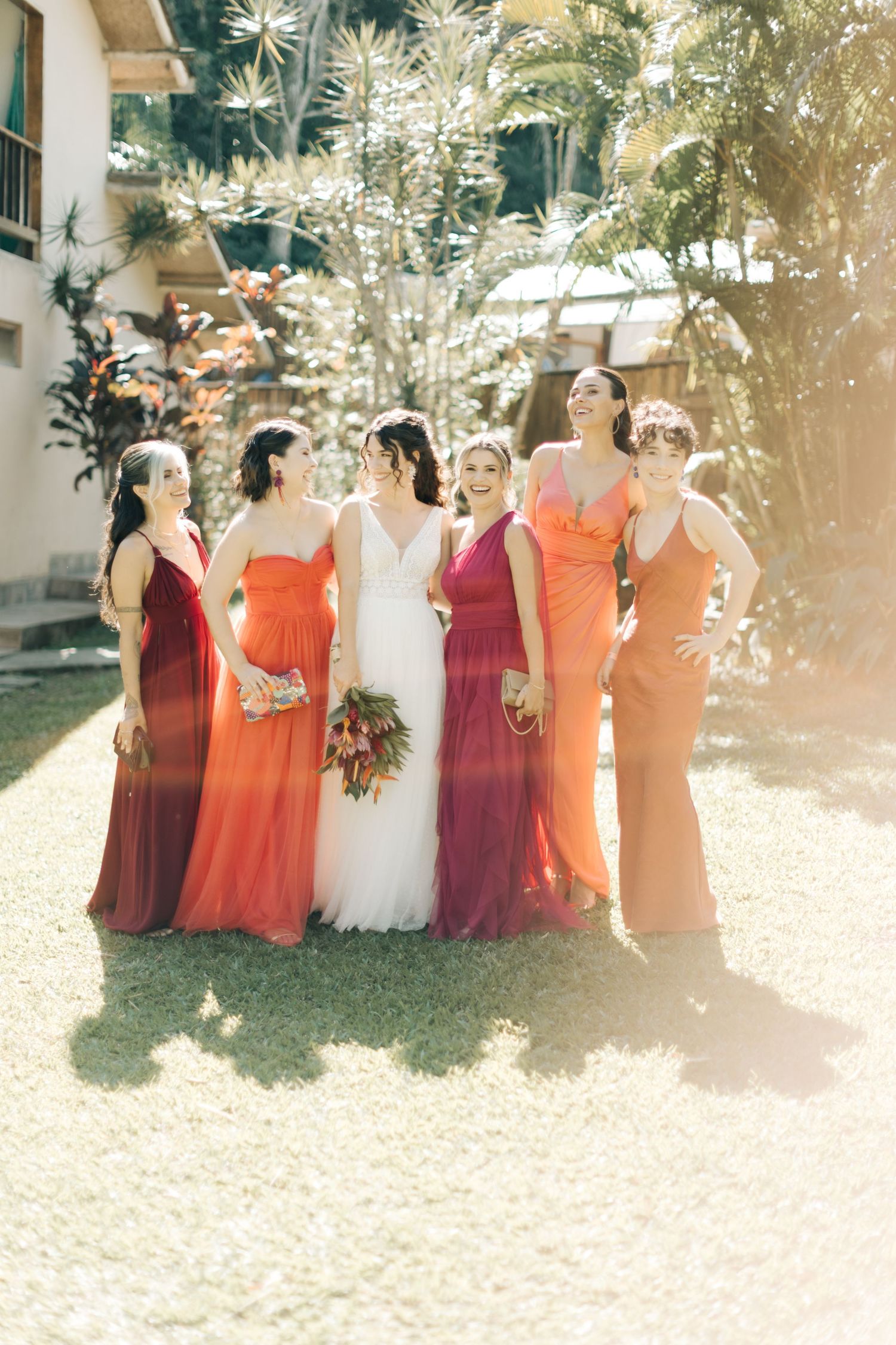 Wedding party wearing burgundy and coral dresses standing together on a lawn with tropical landscaping.