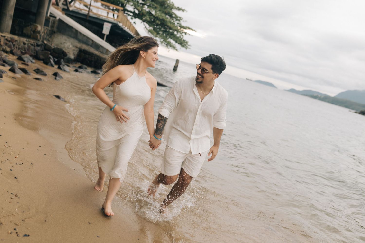 A couple walking hand in hand along the shoreline wearing coordinated white outfits.