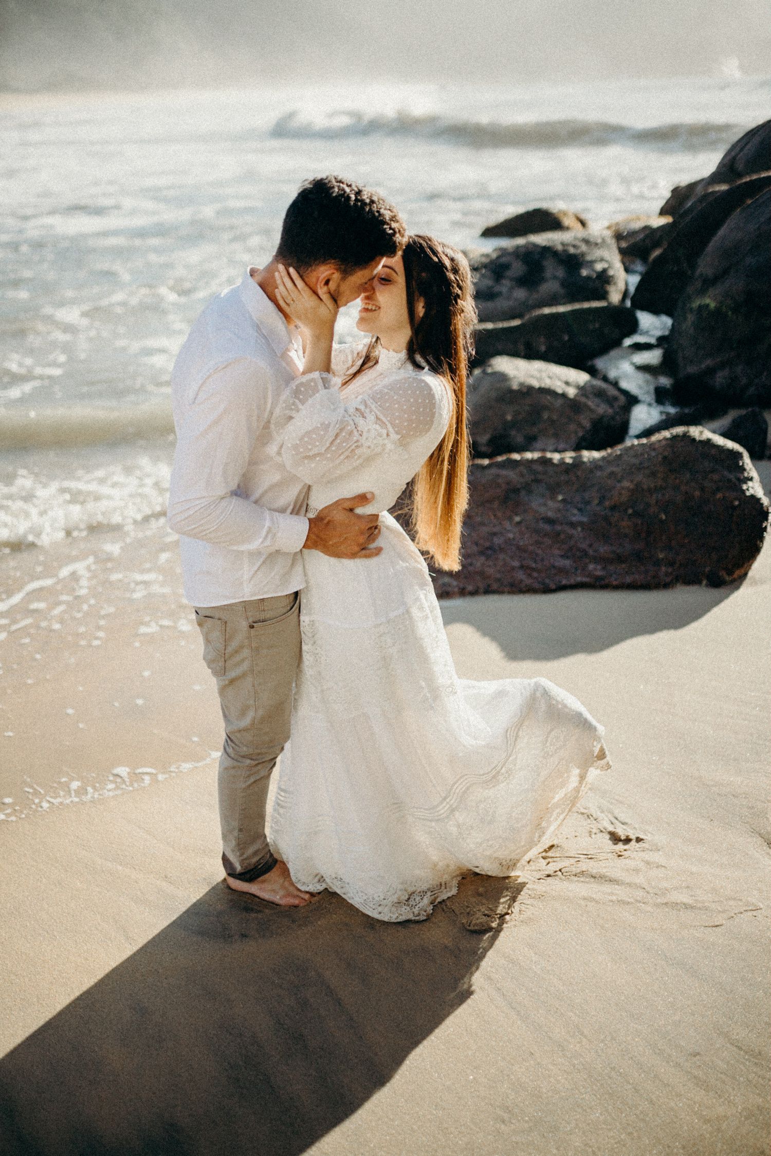 A tender moment between a couple on a beach with waves and volcanic rocks in the background.