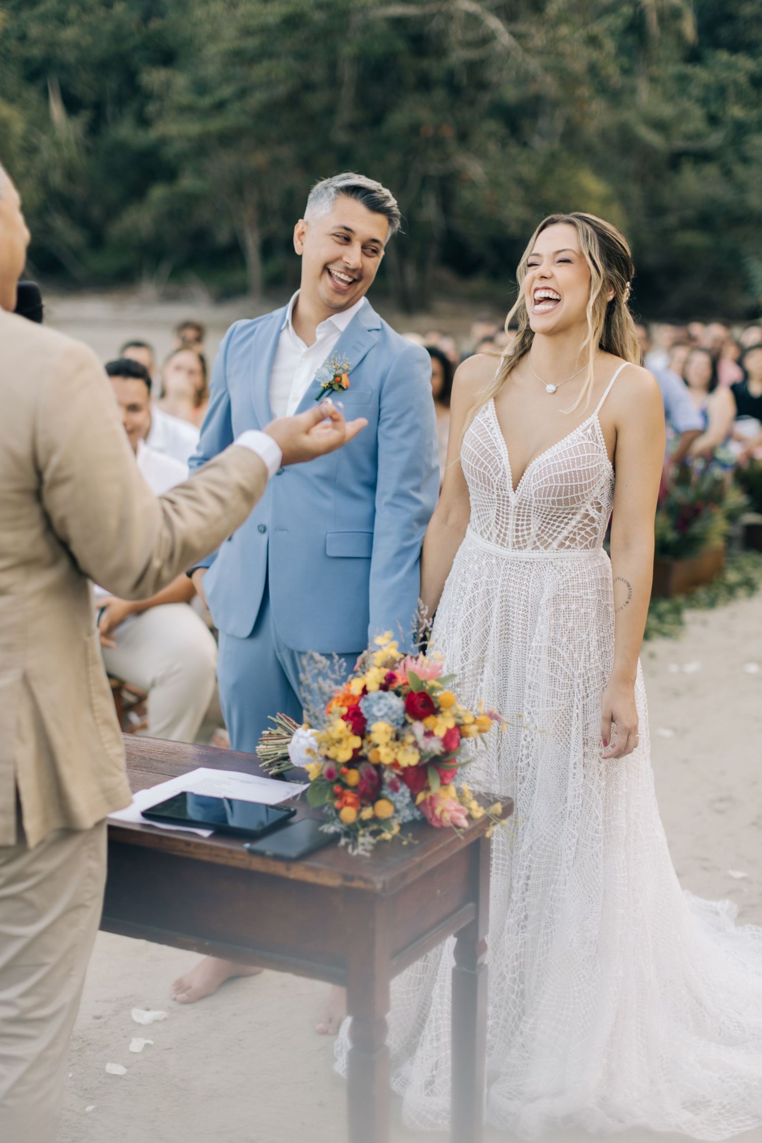 Beach wedding couple laughing with vibrant floral bouquet and rustic decor.