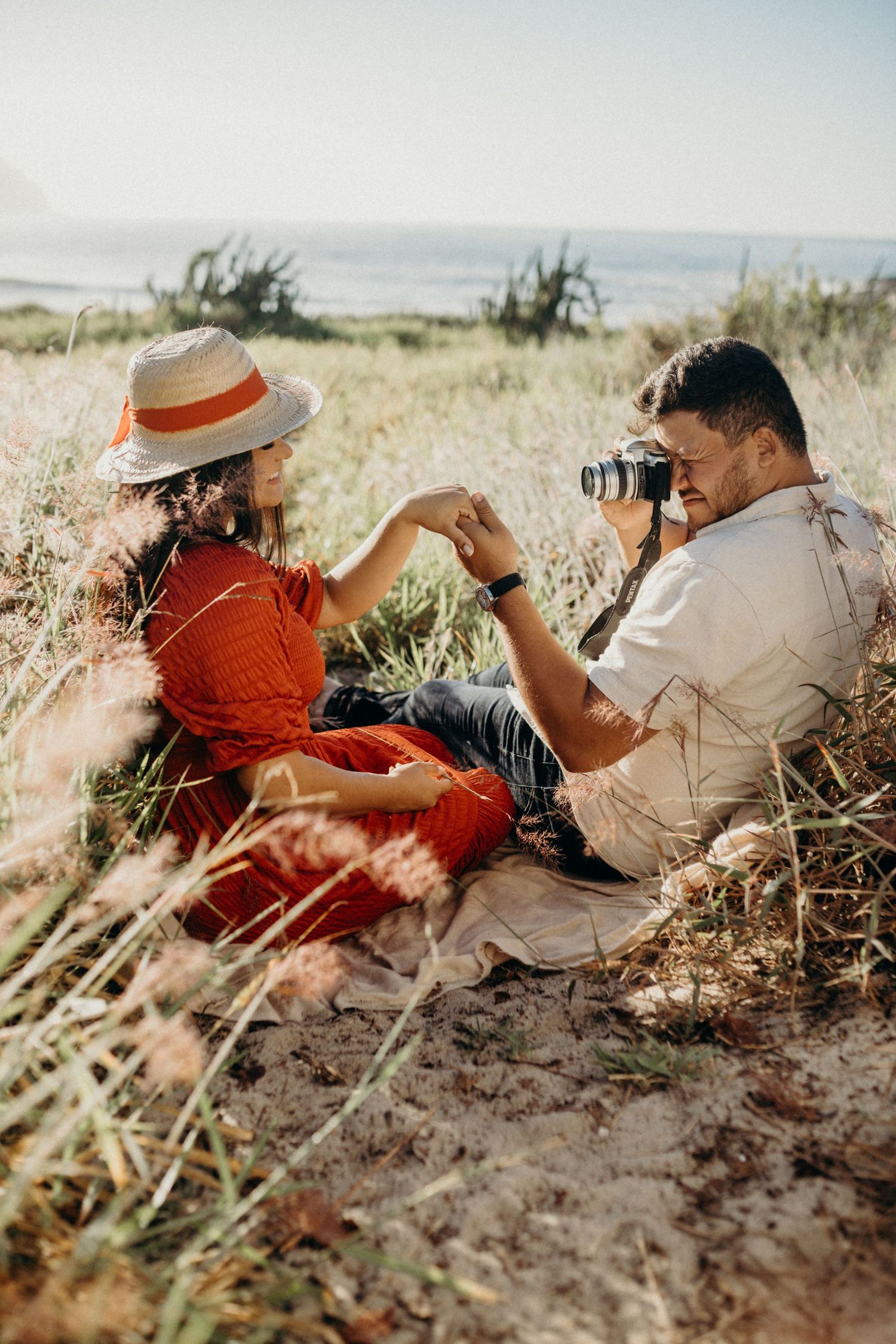 A couple sits in tall grass by the ocean as one takes photos while the other wears a red dress and straw hat.