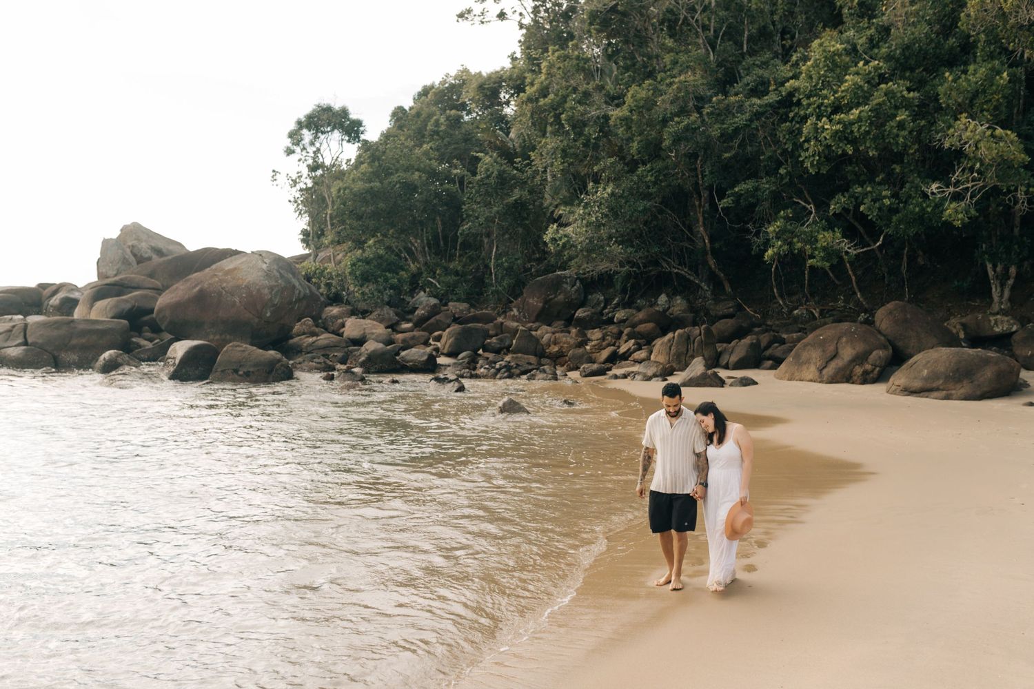 A couple embraces in the ocean waves near a rocky beach at sunset.