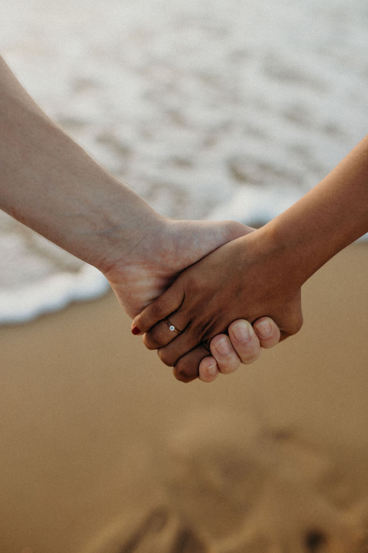 Close-up of two people holding hands on a sandy beach, with one person wearing an engagement ring.
