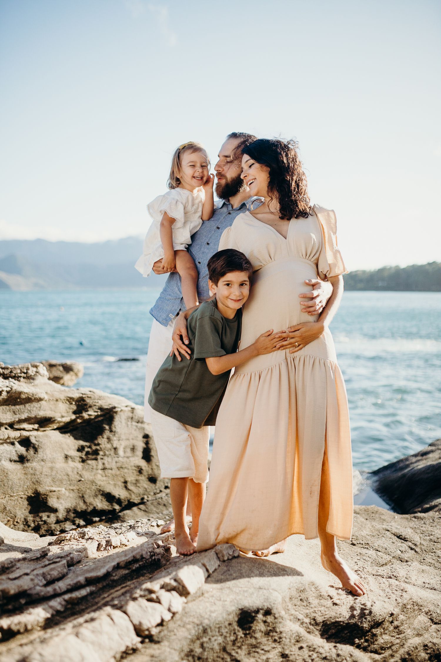 Happy family poses together on rocky beach with ocean waves in background.
