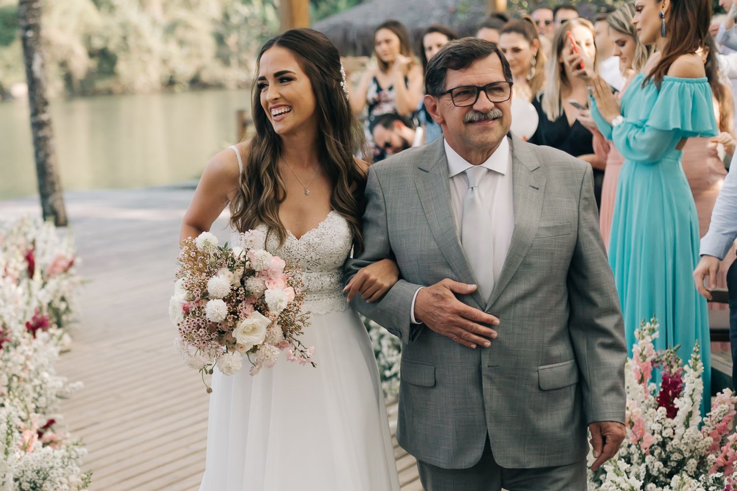 Wedding processional on wooden deck with pink and white floral arrangements and guests watching.