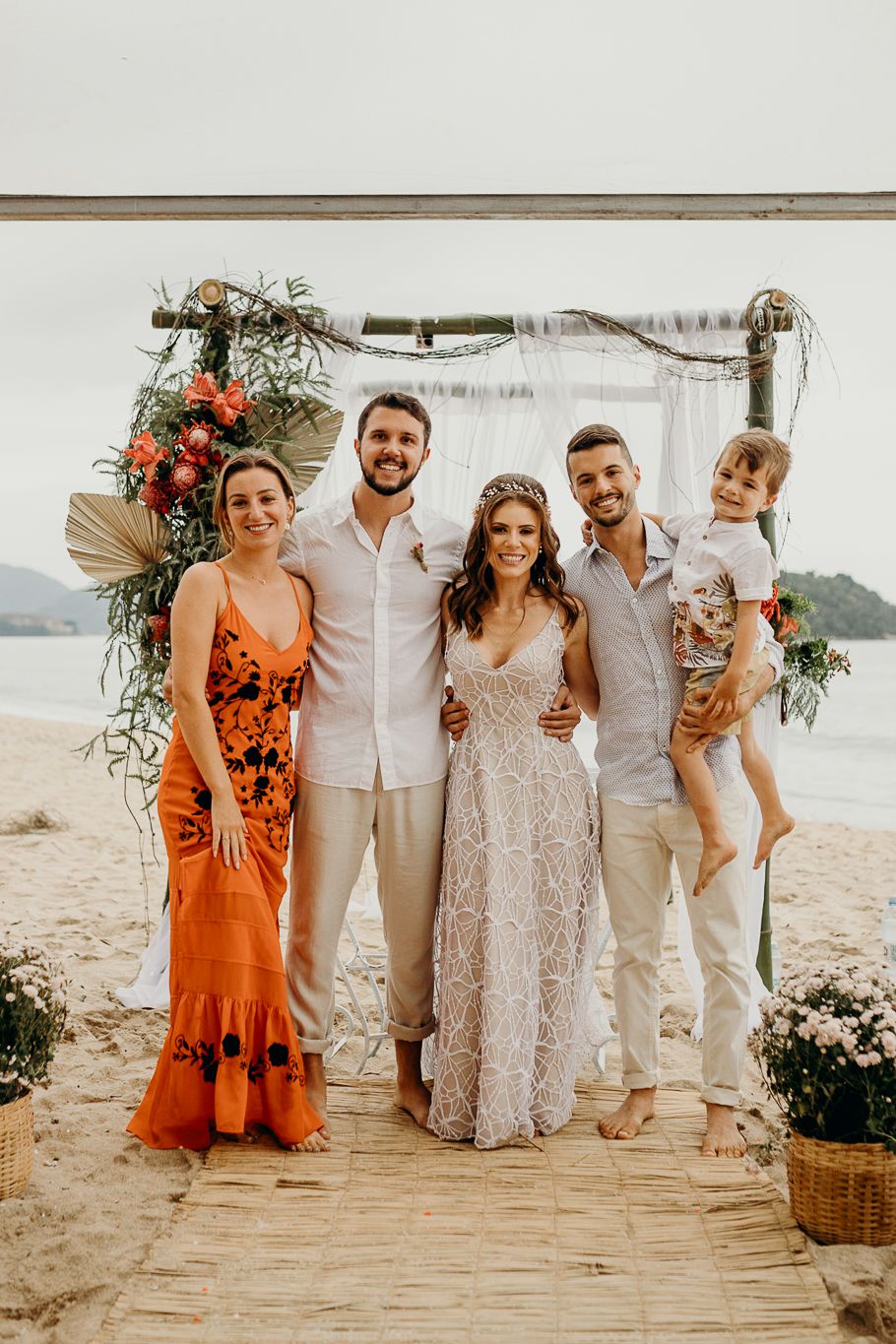 A beachside wedding party poses under a decorated arch with mountains in the background at sunset.