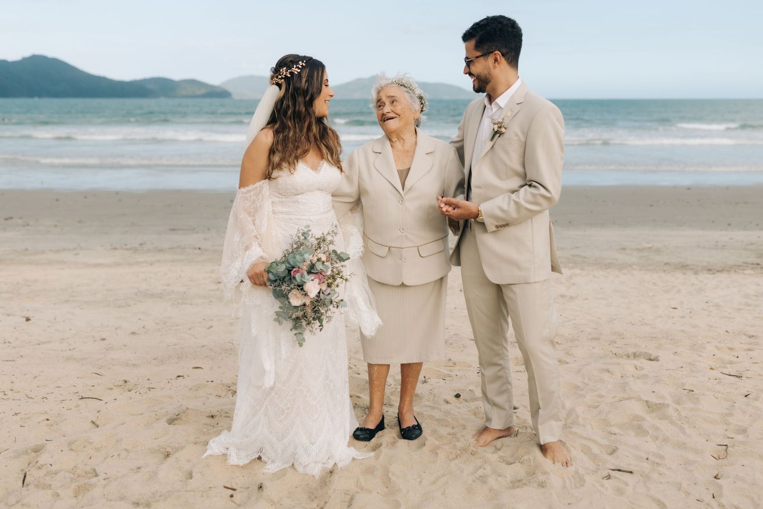Wedding party members share special moment on sandy beach with mountains in background.
