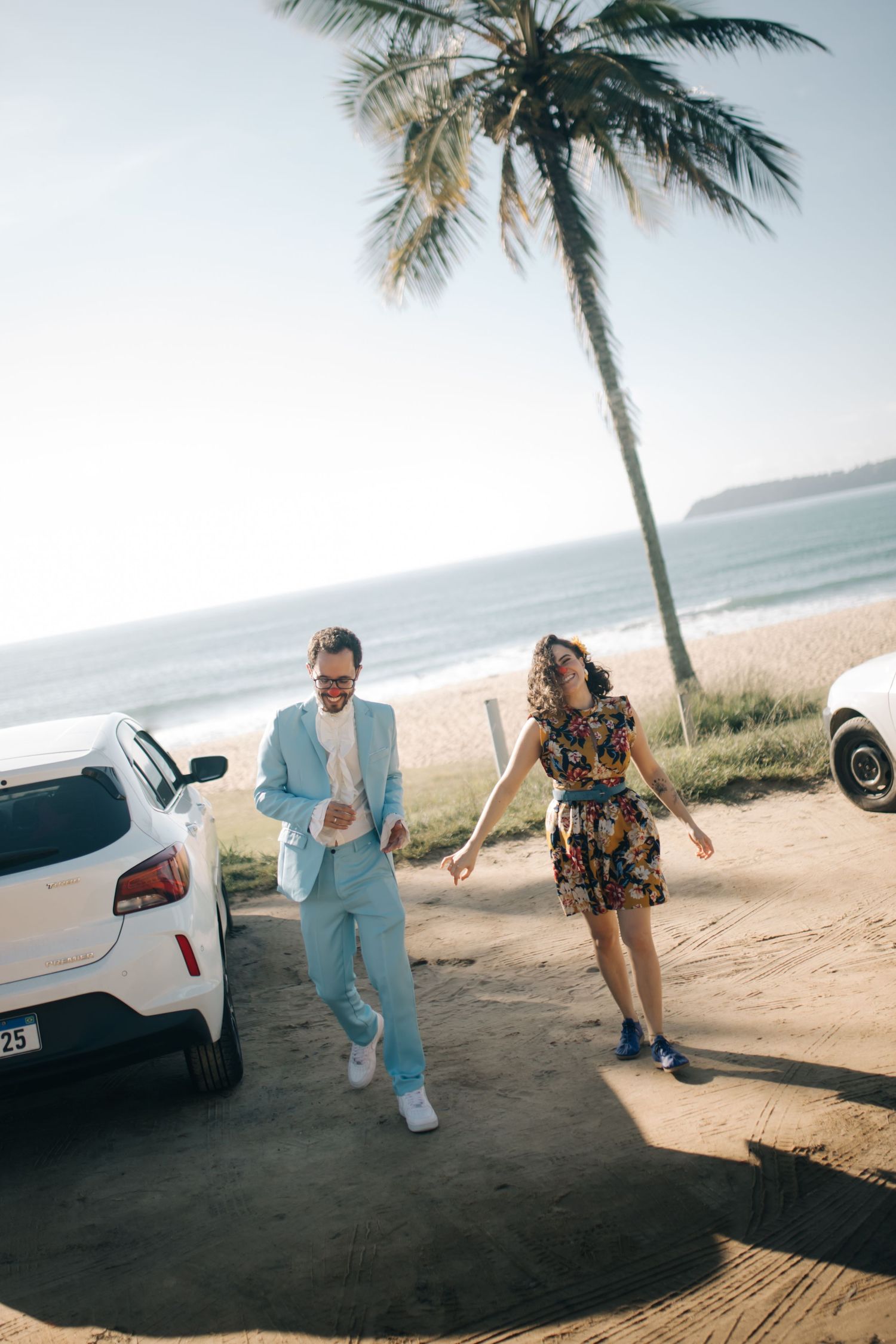 Playful couple in summer outfits dancing by car on palm-lined beach.