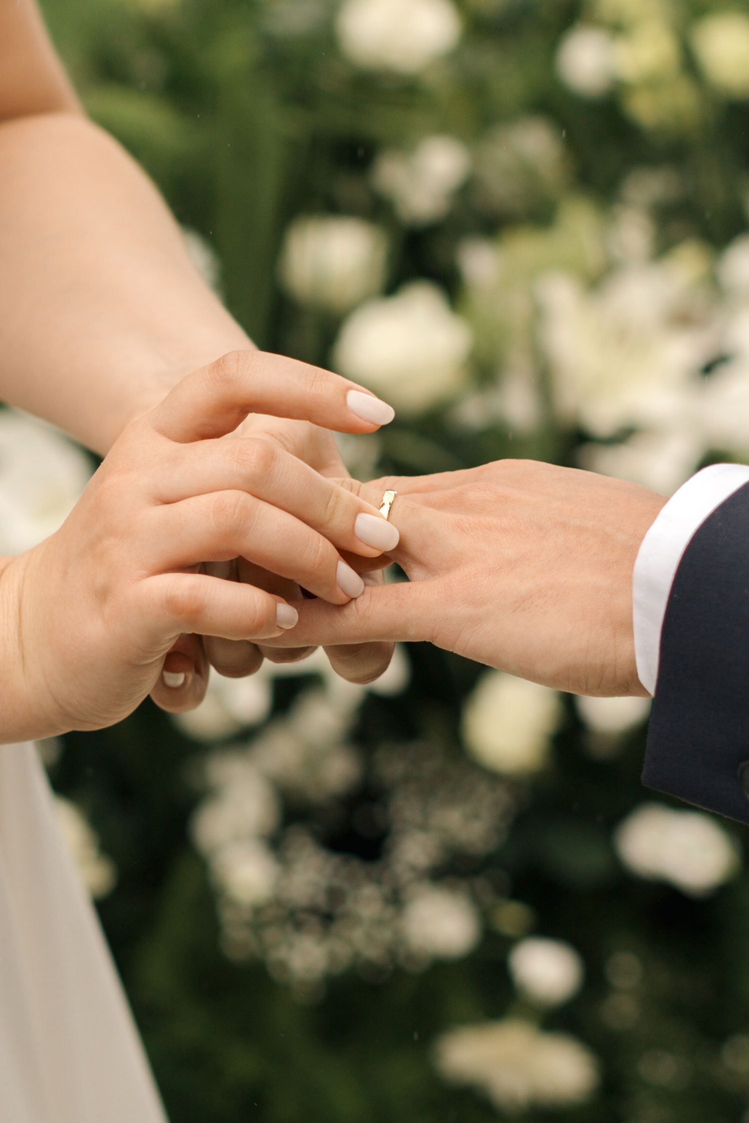 Close-up of hands exchanging wedding rings against a backdrop of white flowers.